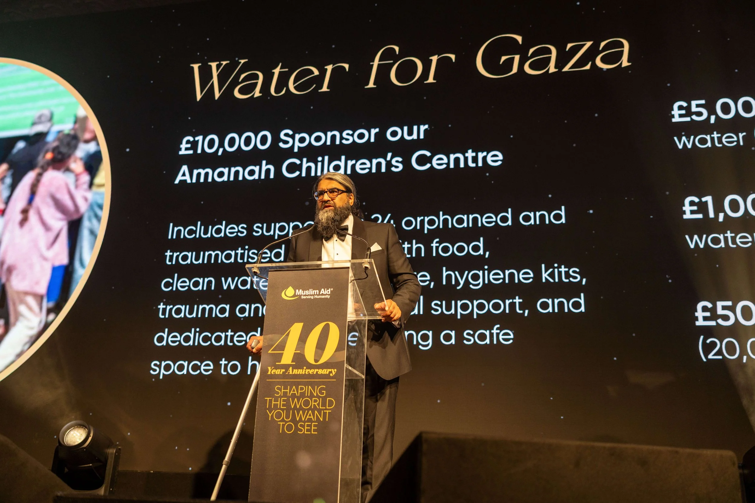 A man in a suit with glasses and a beard giving a speech at a charity event for Muslim Aid, standing behind a podium with a poster celebrating the 40th anniversary of the organization. The large screen behind him displays information about a water do