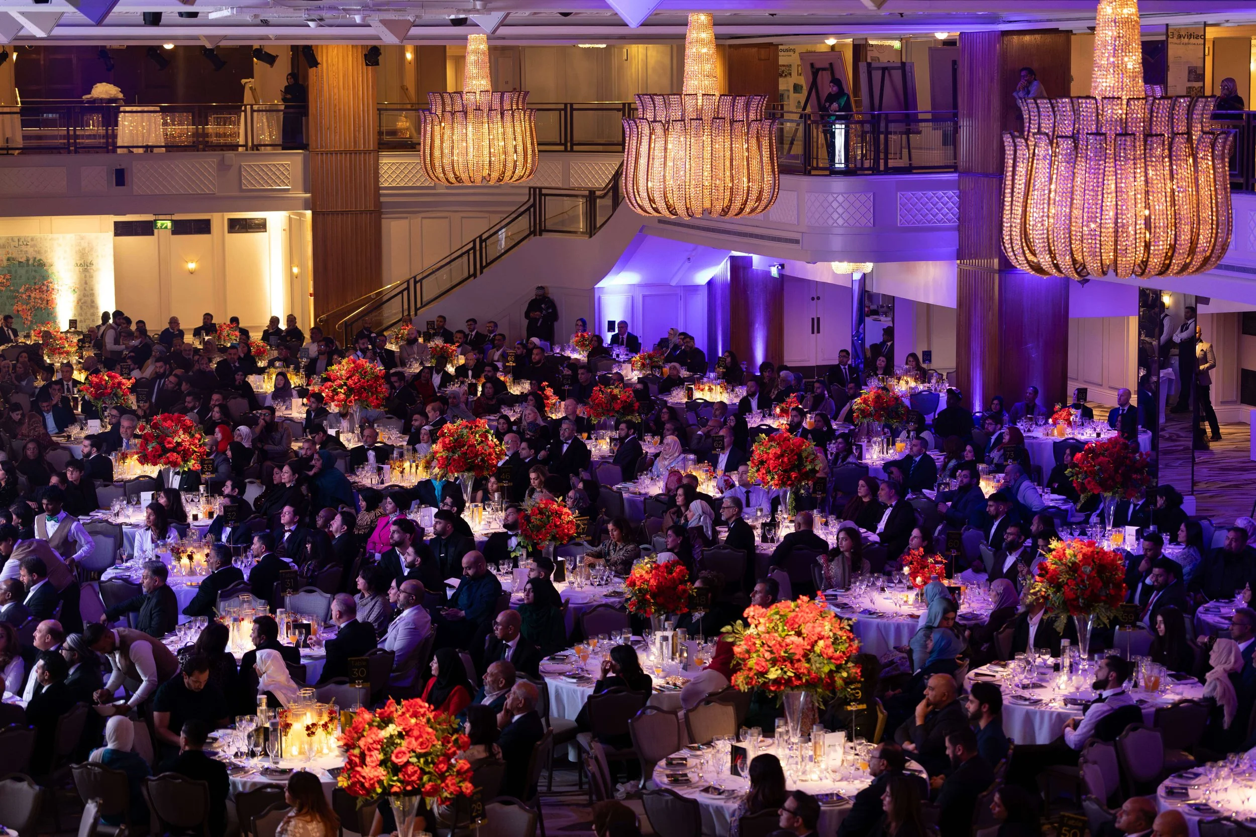 A large banquet hall filled with round tables decorated with tall floral centerpieces, chandeliers, and purple lighting, hosting many formally dressed guests at a formal event or conference.