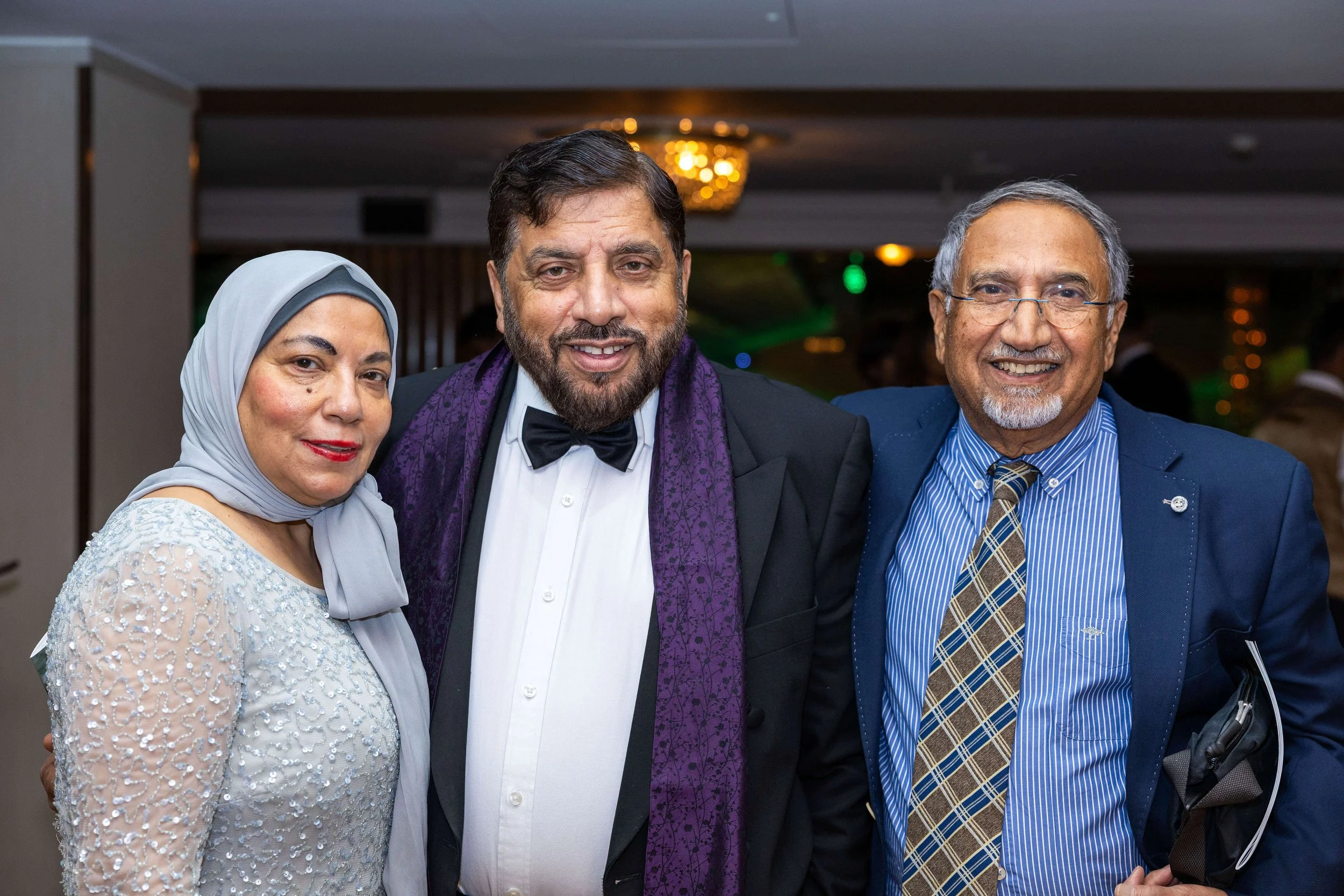 Three people dressed in formal attire at an event, smiling at the camera, with a background of a dimly lit room with green and yellow lights.