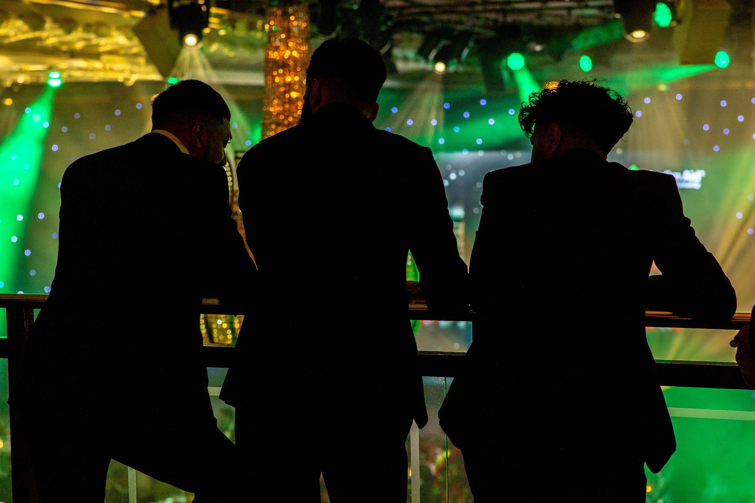 Silhouettes of three men in suits standing at a bar, with colorful stage lighting and sparkling decorations in the background.
