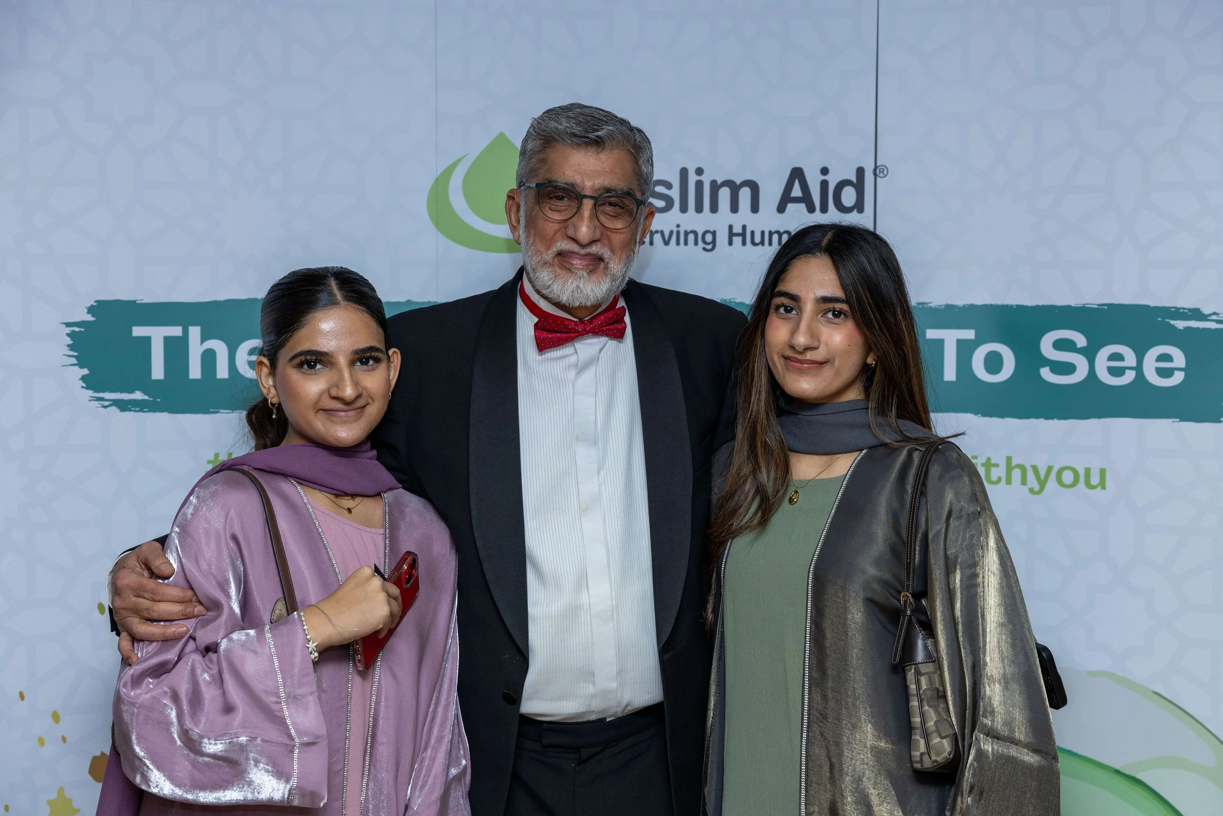 Three people standing close together at a charity event for Slim Aid, with a man in the center wearing a black suit, white shirt, and red bow tie, and two women wearing traditional South Asian attire, one in a purple outfit and the other in a green o