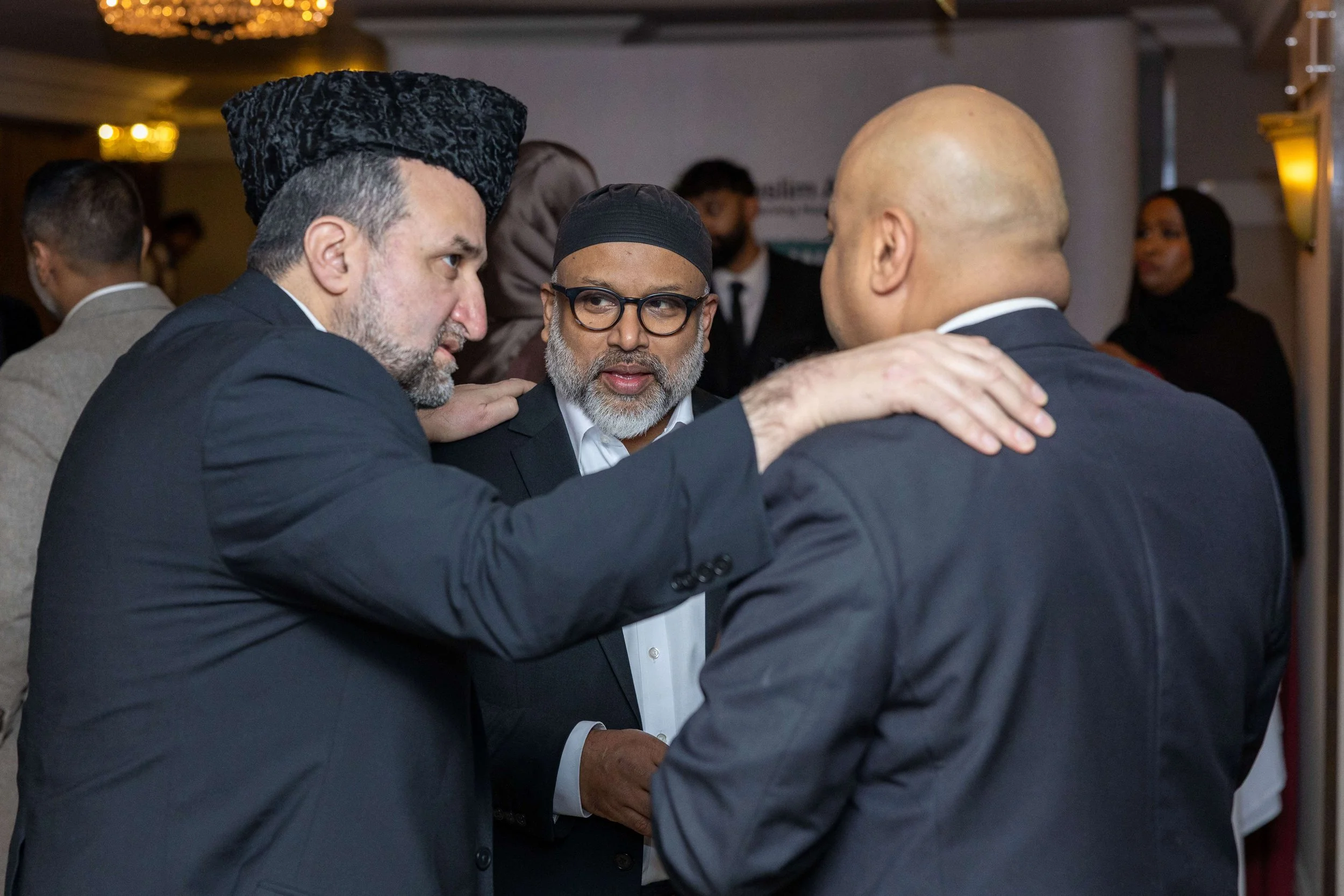 Three men in suits engaged in a serious conversation at an indoor event, with other people in the background.