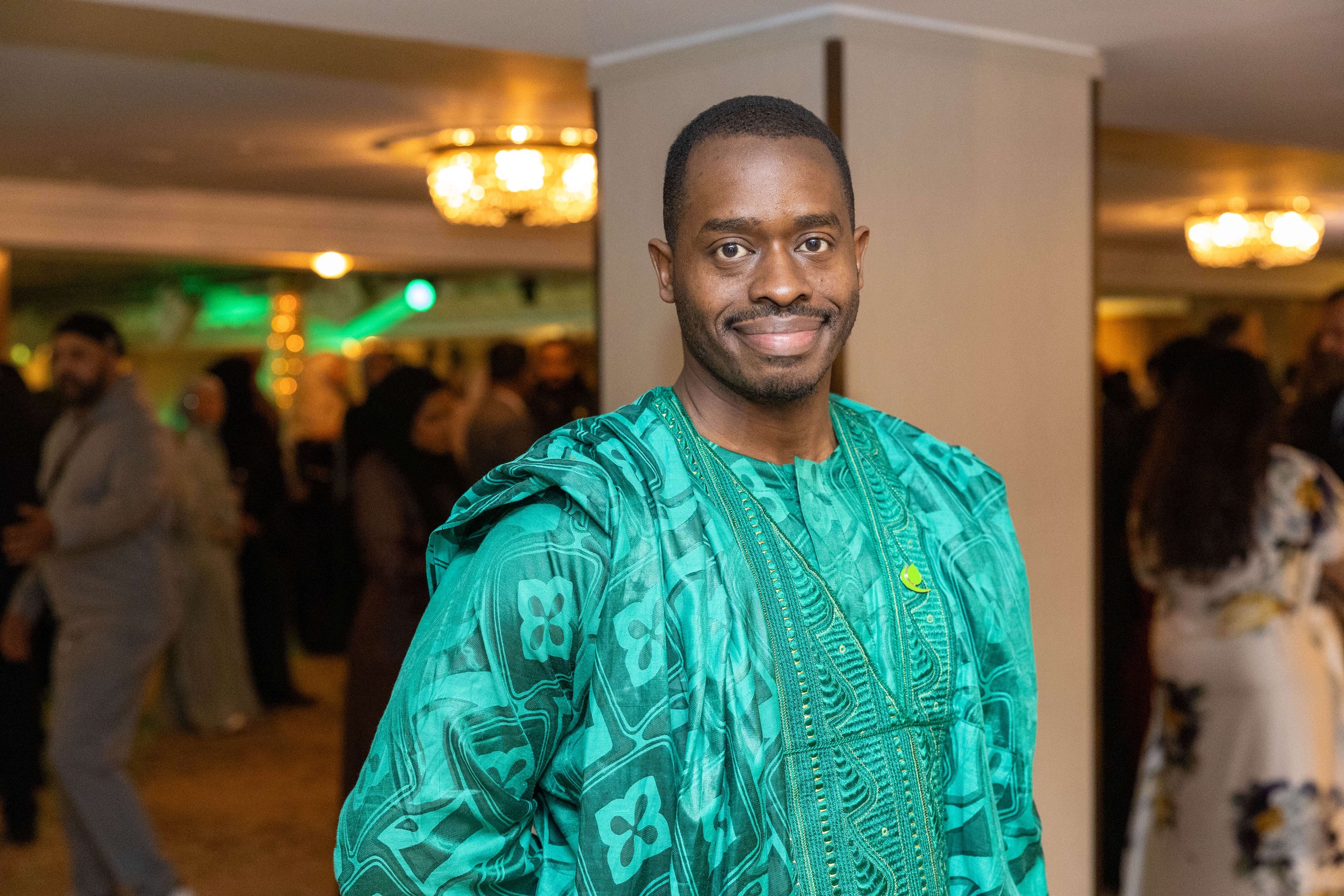 A smiling man in a traditional African outfit standing at a social gathering or event, with people and decorations in the background.