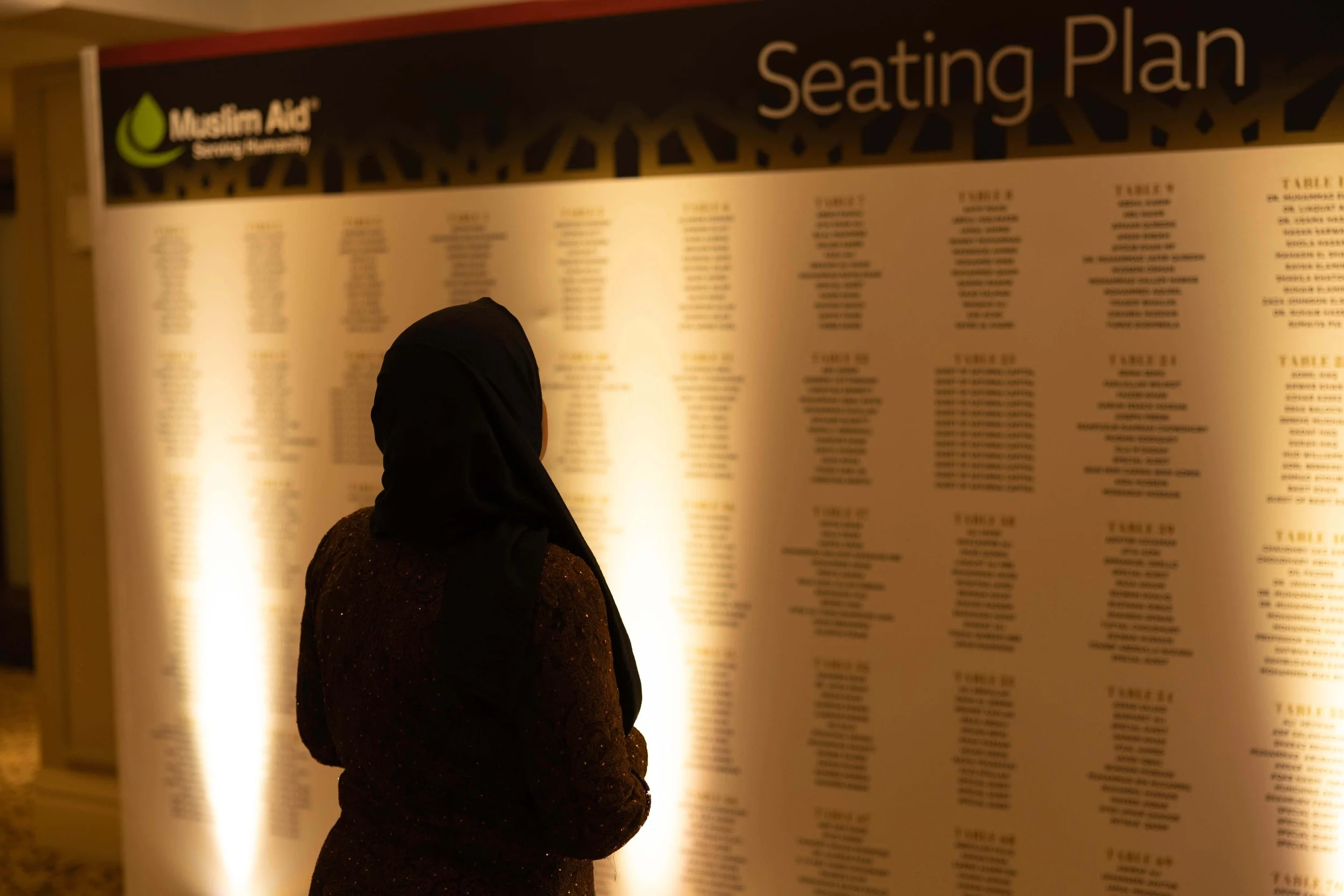 A person wearing a black hijab and a dark sweater looking at a large, illuminated seating plan board at a Muslim Add Serving Sanctuary event.