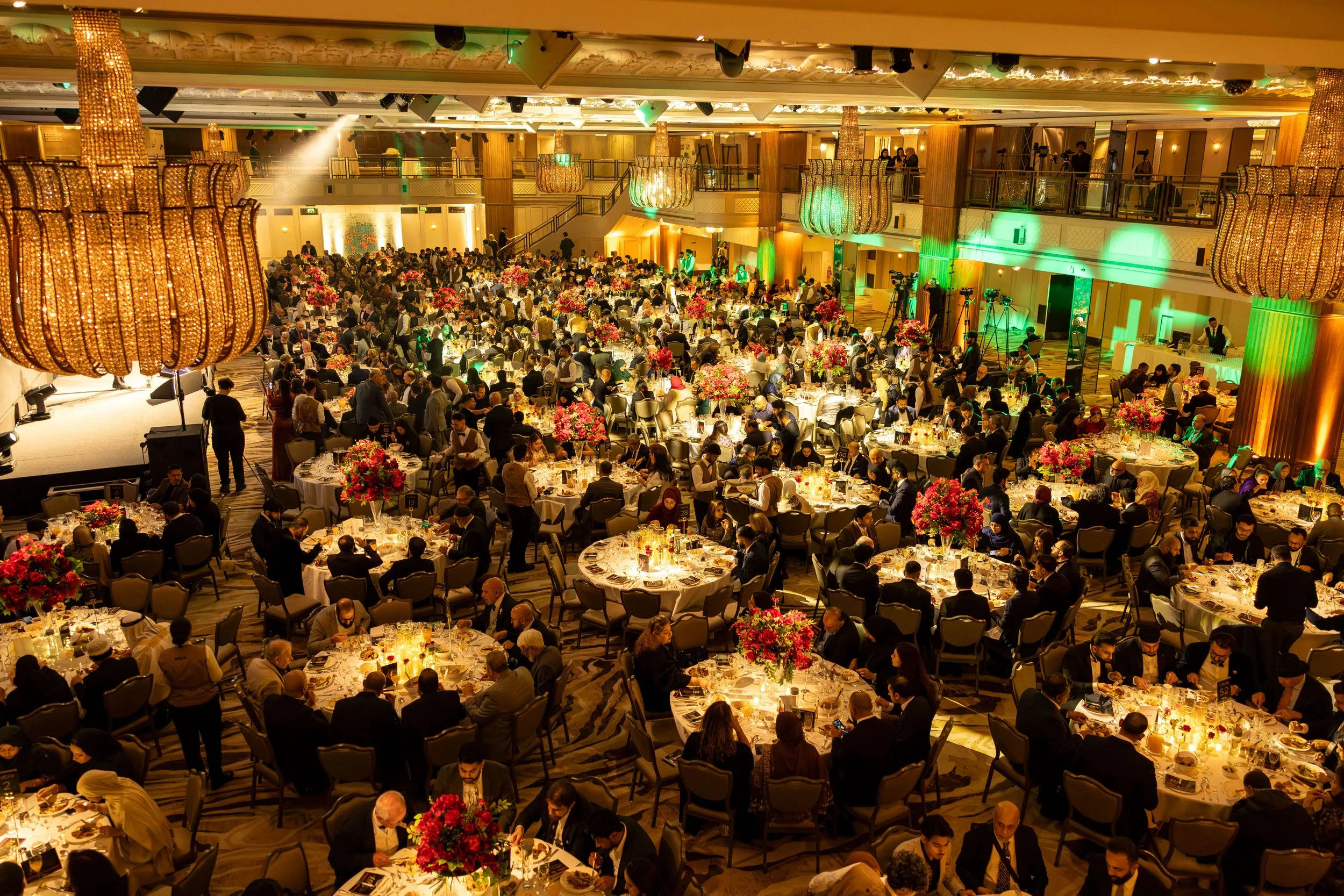 A large banquet hall filled with round tables decorated with pink and red flower centerpieces, elegantly dressed guests, and a stage with lighting, chandeliers, and a balcony area overhead.