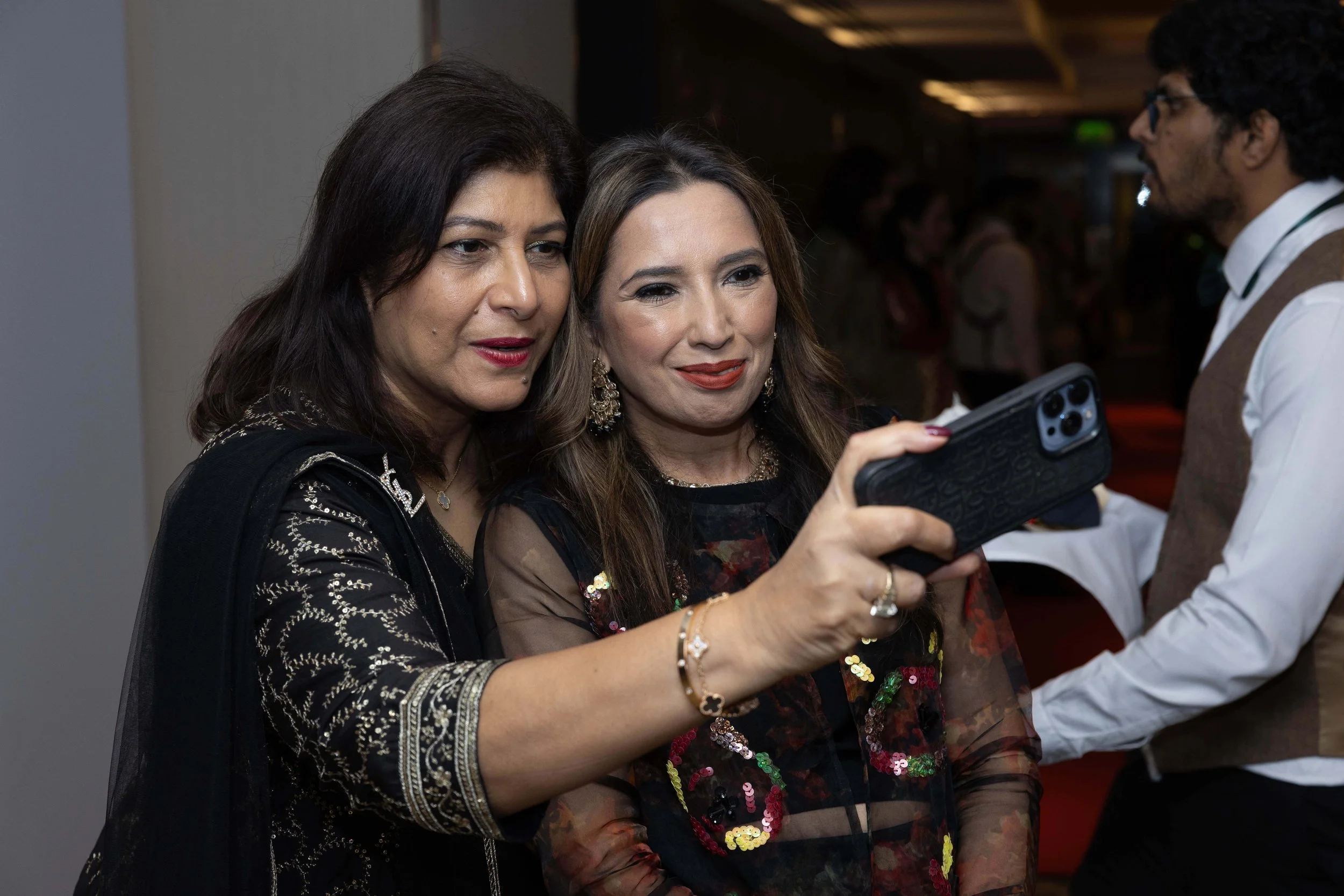 Two women taking a selfie together at an indoor event, with a man standing nearby in the background.
