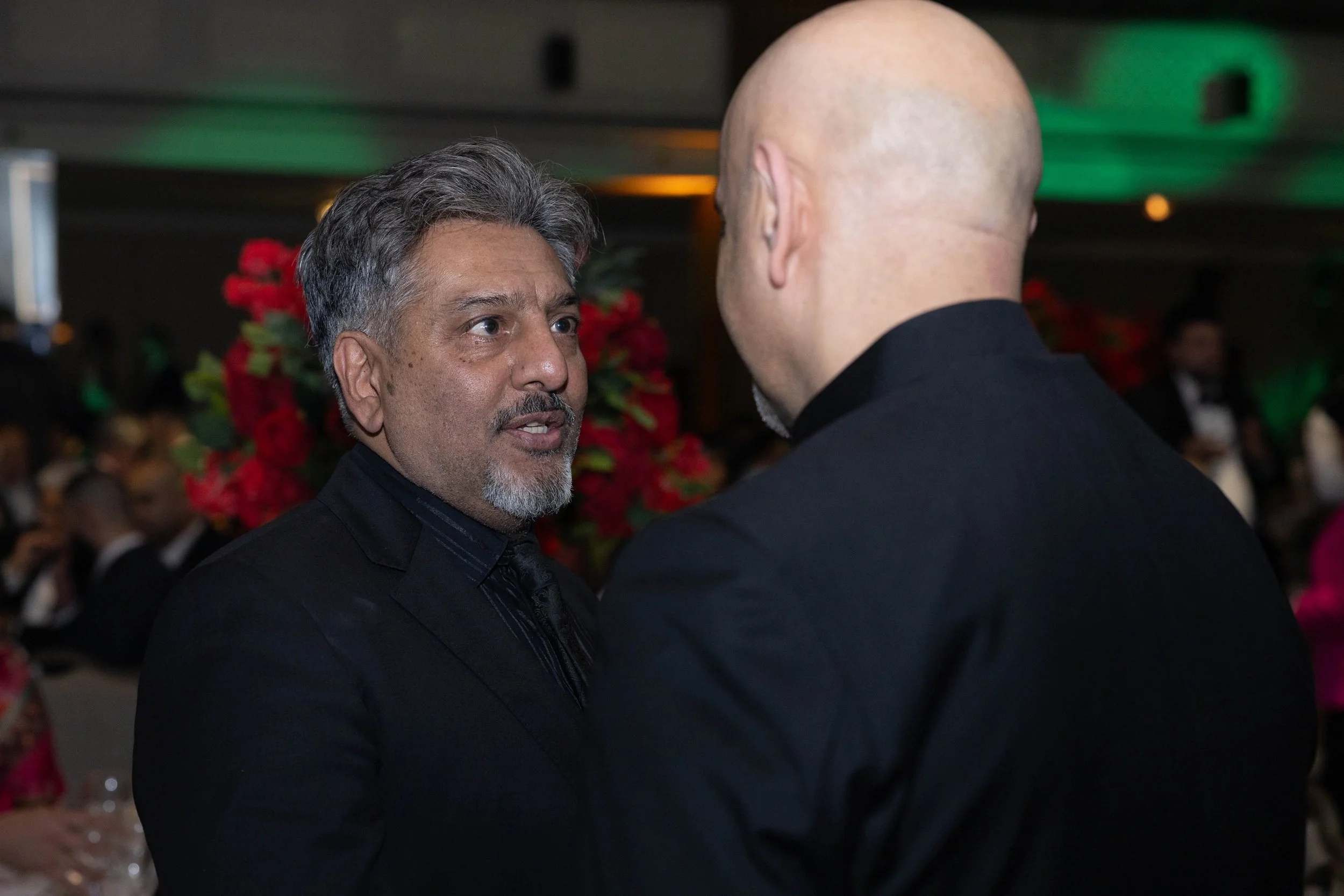 Two men in formal attire engaging in conversation at an indoor event with a blurred background of other guests and red floral decorations.