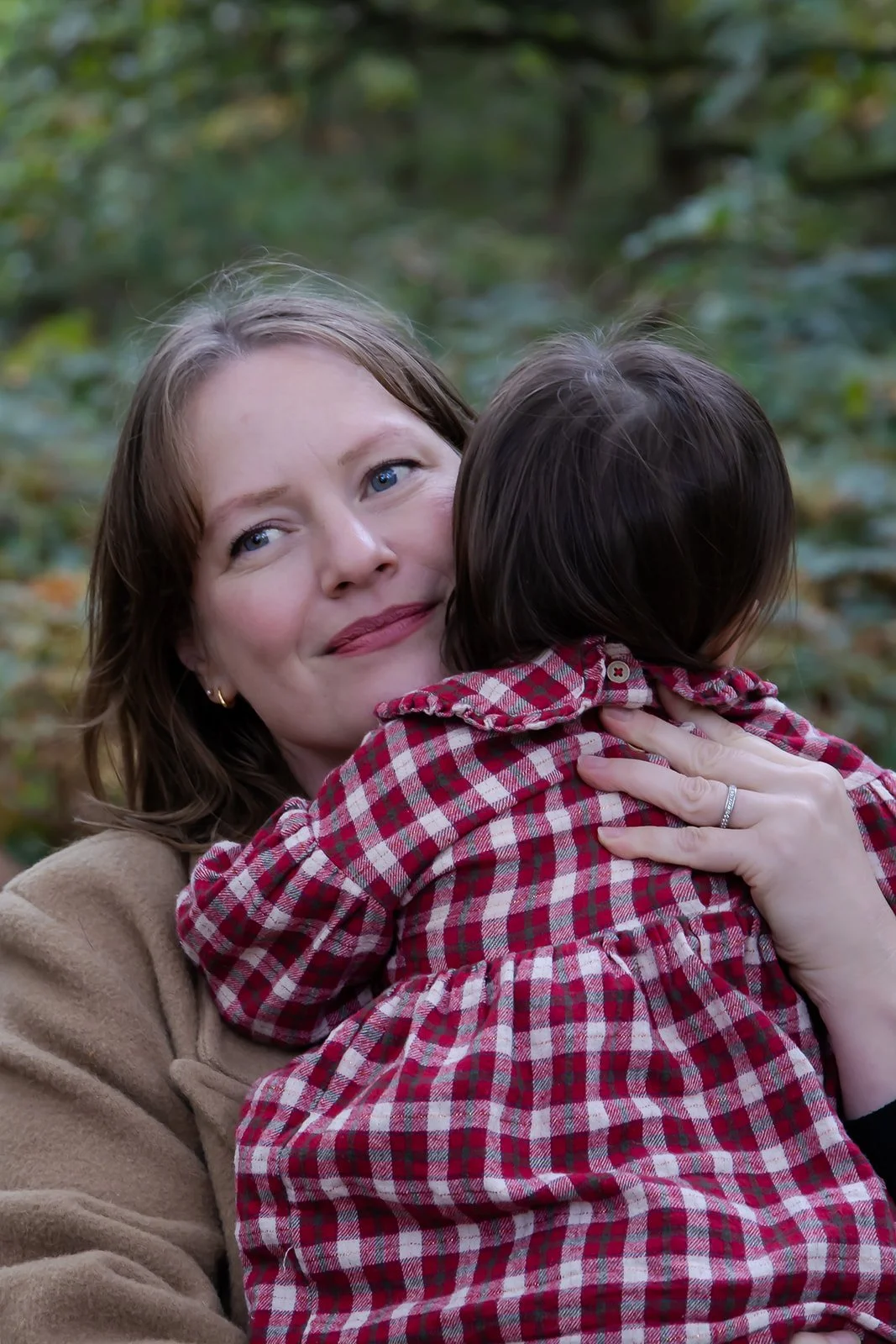A woman with brown hair and blue eyes hugging a child with dark hair, wearing a red and black plaid shirt, in an outdoor setting with green foliage in the background.