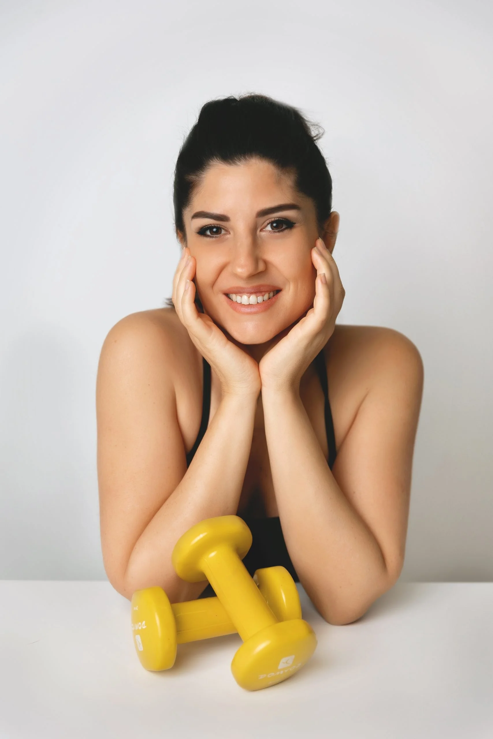 Una donna sorridente con i capelli raccolti, poggiando il viso sulle mani, di fronte a due manubri gialli sul tavolo.