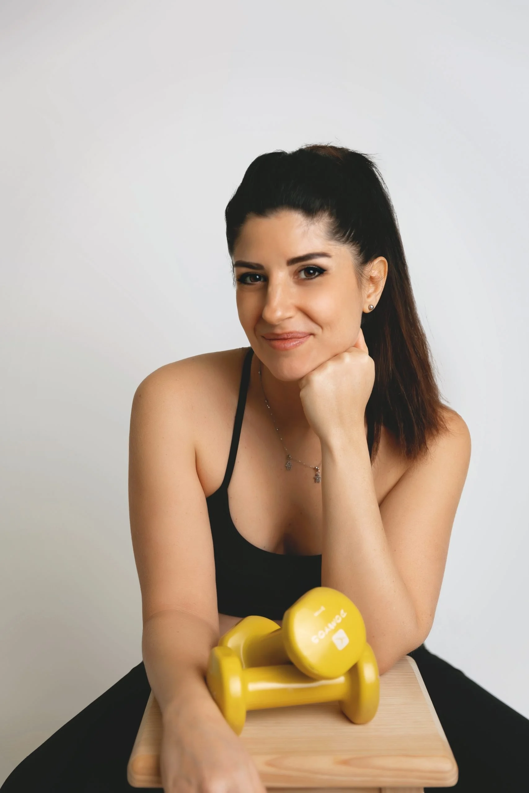 Una donna sorridente con capelli castani e abbigliamento nero, che si appoggia sulla mano destra, con due manubri gialli posizionati su un tavolo di legno davanti a lei, in uno sfondo bianco.