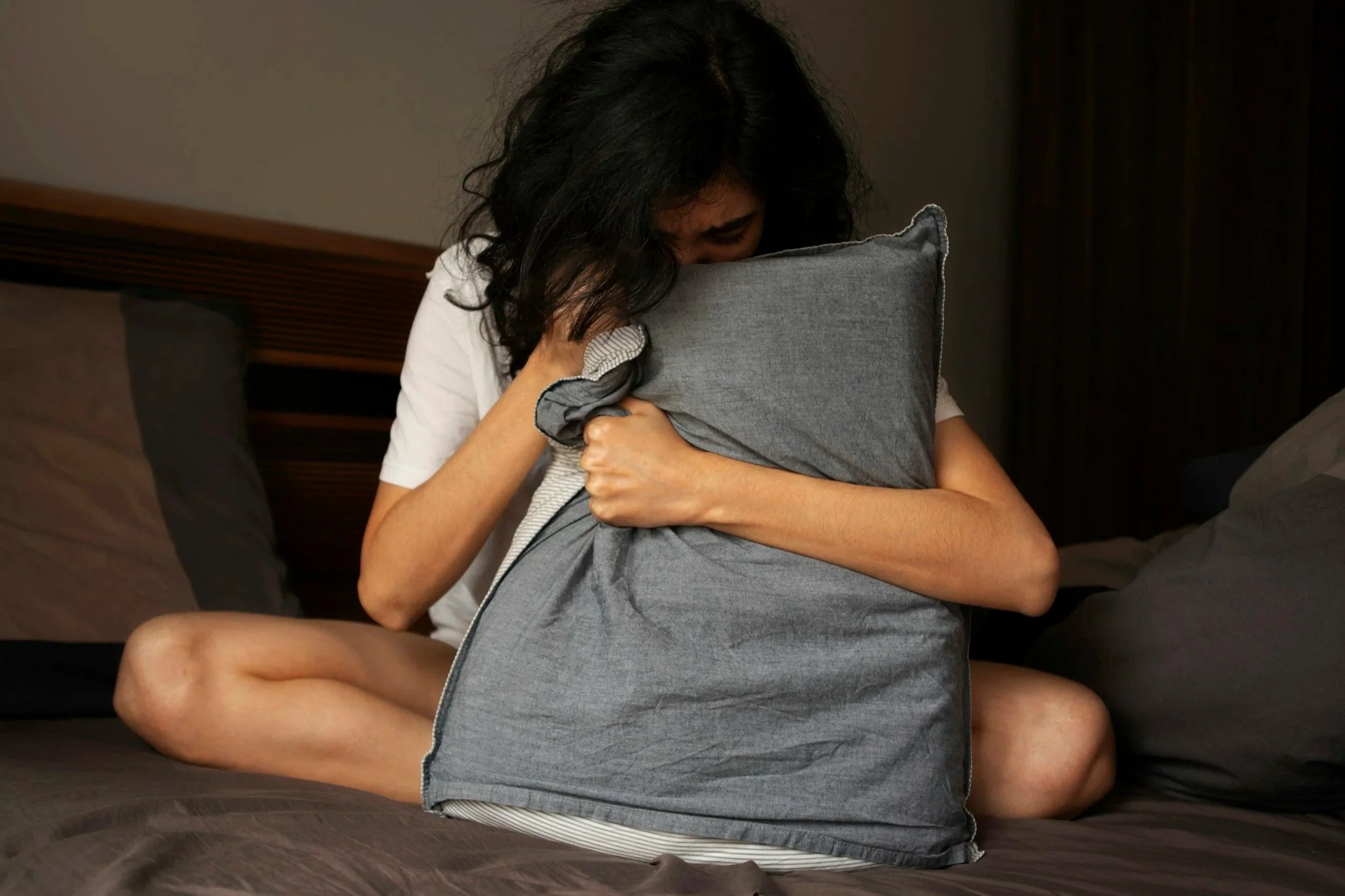 A woman sitting on a bed holding a pillow to her stomach, illustrating chronic pelvic pain, endometriosis support, and pelvic girdle relief.