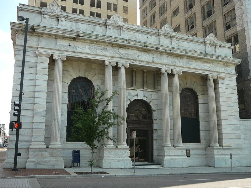 The front of the historic Marble Bank building. White concrete columns and large arched entryway and windows.