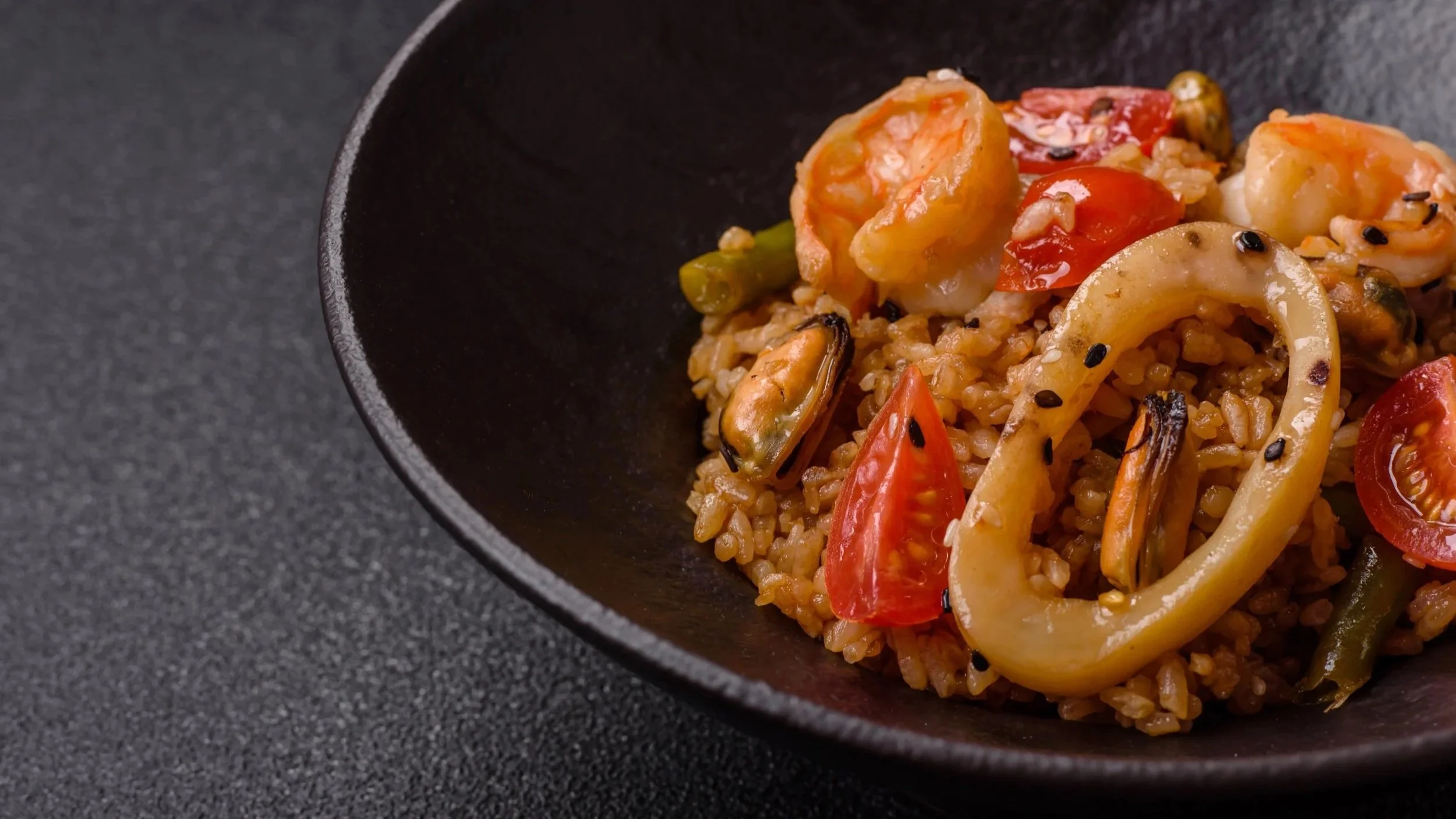 Close-up of a bowl of fried rice with shrimp, cherry tomatoes, squid, and black sesame seeds.