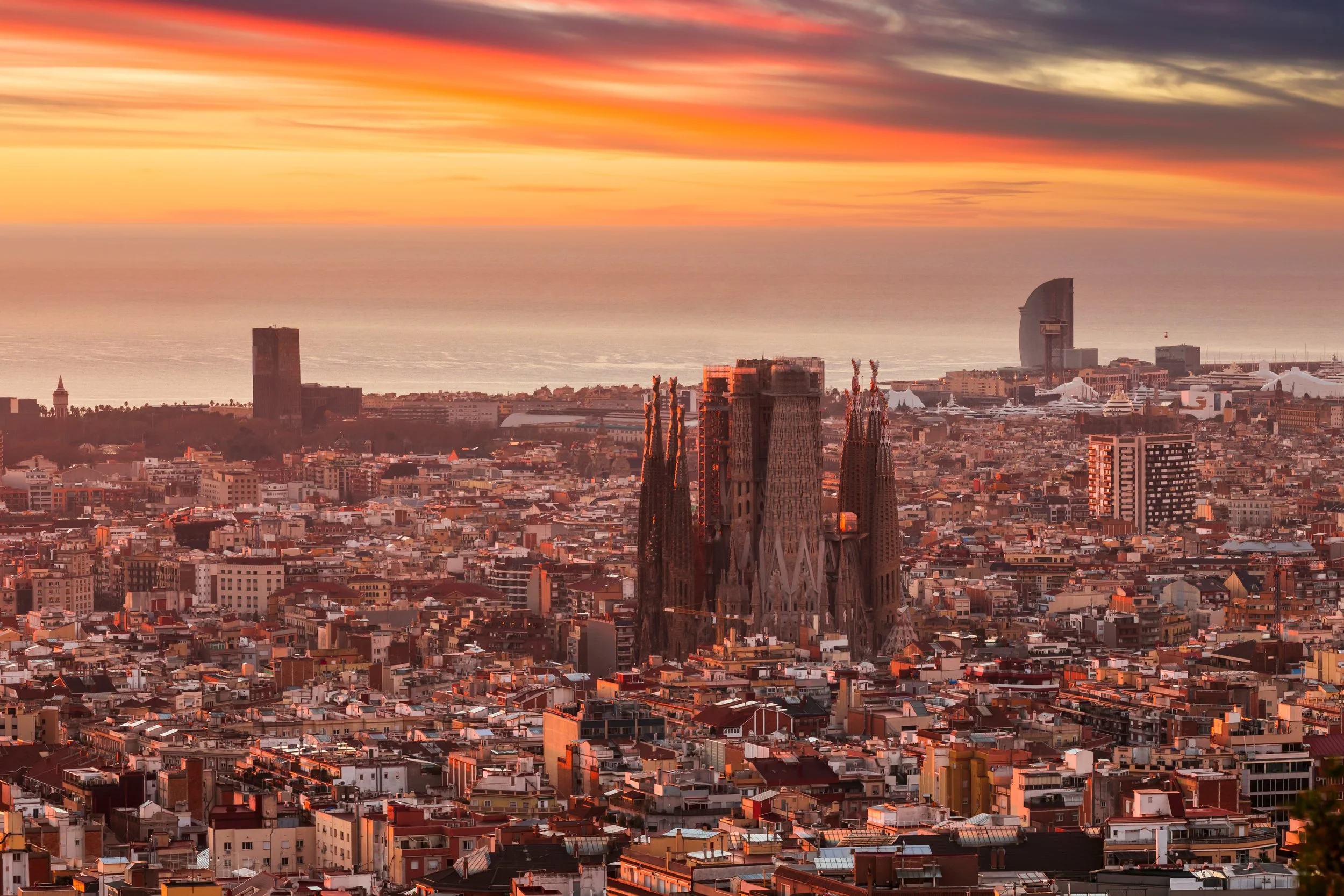 Aerial view of Barcelona cityscape at sunset, featuring the Sagrada Família basilica in the foreground and the coastline in the background.