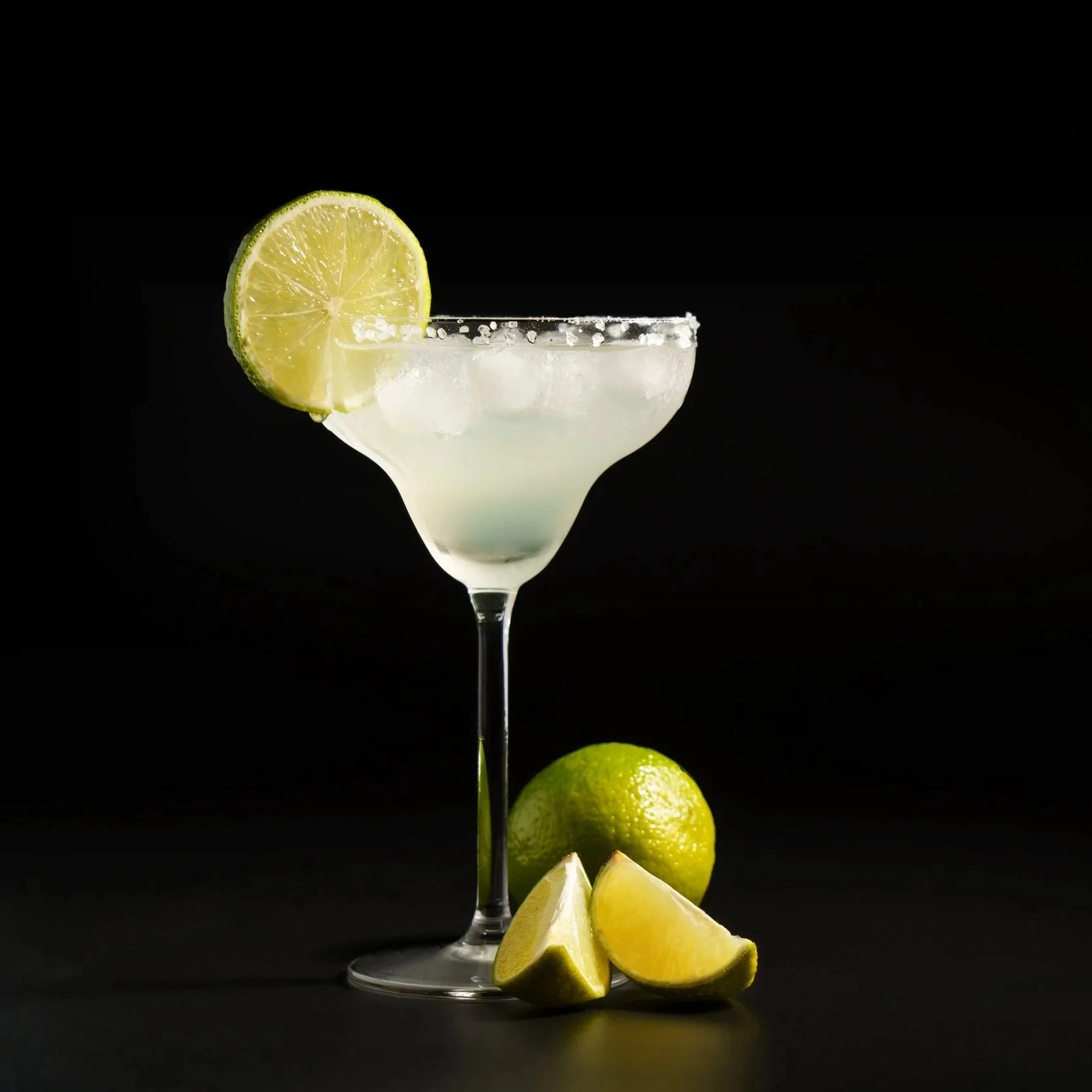 A cocktail in a classic margarita glass with a salted rim and a lime wedge garnish, placed beside a whole lime and lime wedges, against a black background.