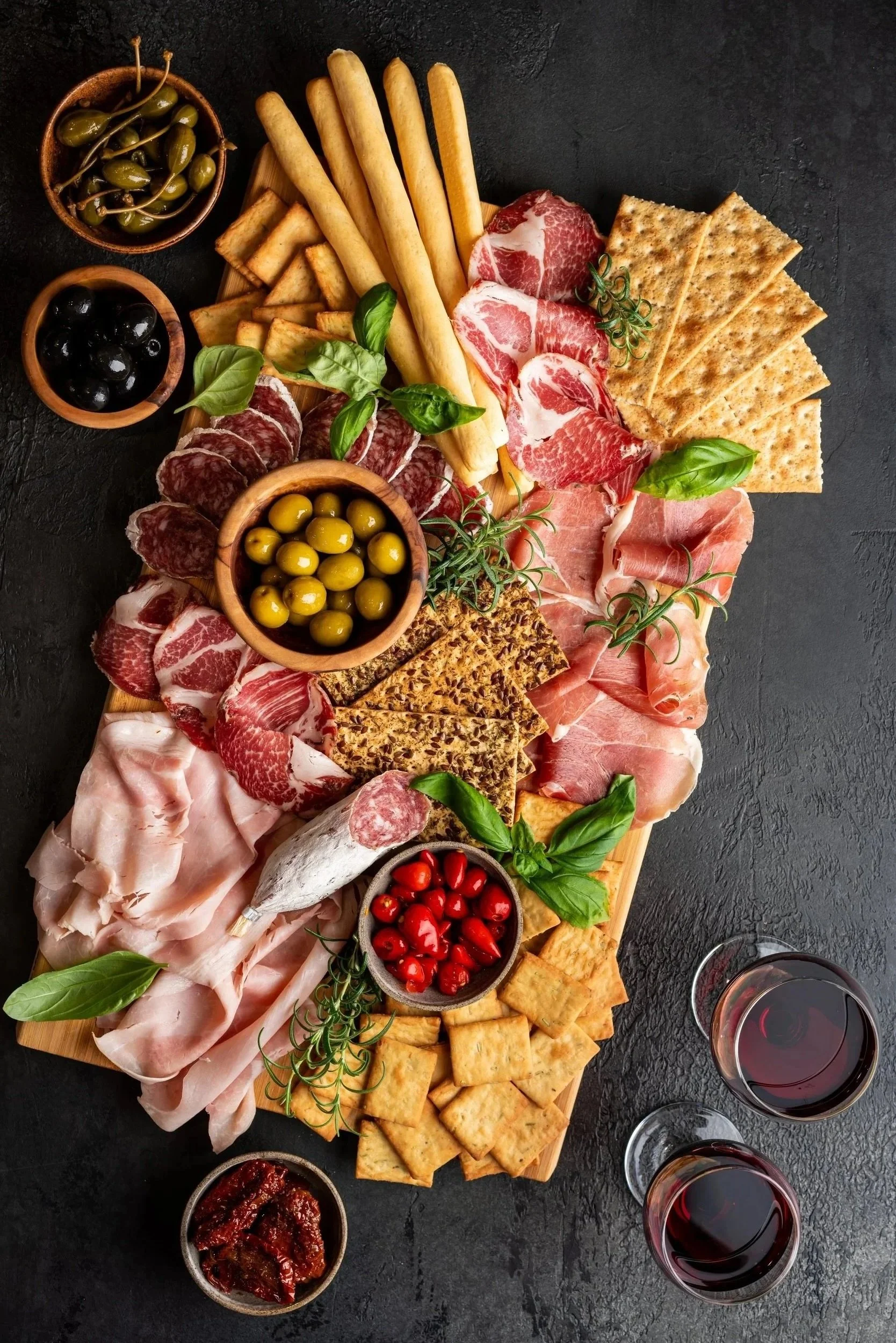 Charcuterie board with assorted cured meats, cheeses, crackers, breadsticks, and bowls of olives, cherry tomatoes, capers, grape, and sun-dried tomatoes, with glasses of red wine.