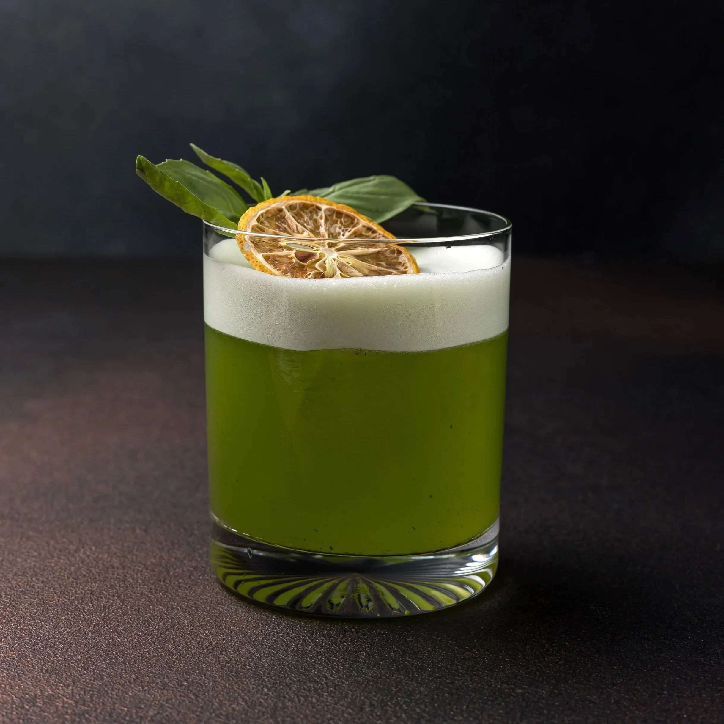 A glass of green cocktail garnished with dried lemon slices and fresh green leaves, set against a dark background.