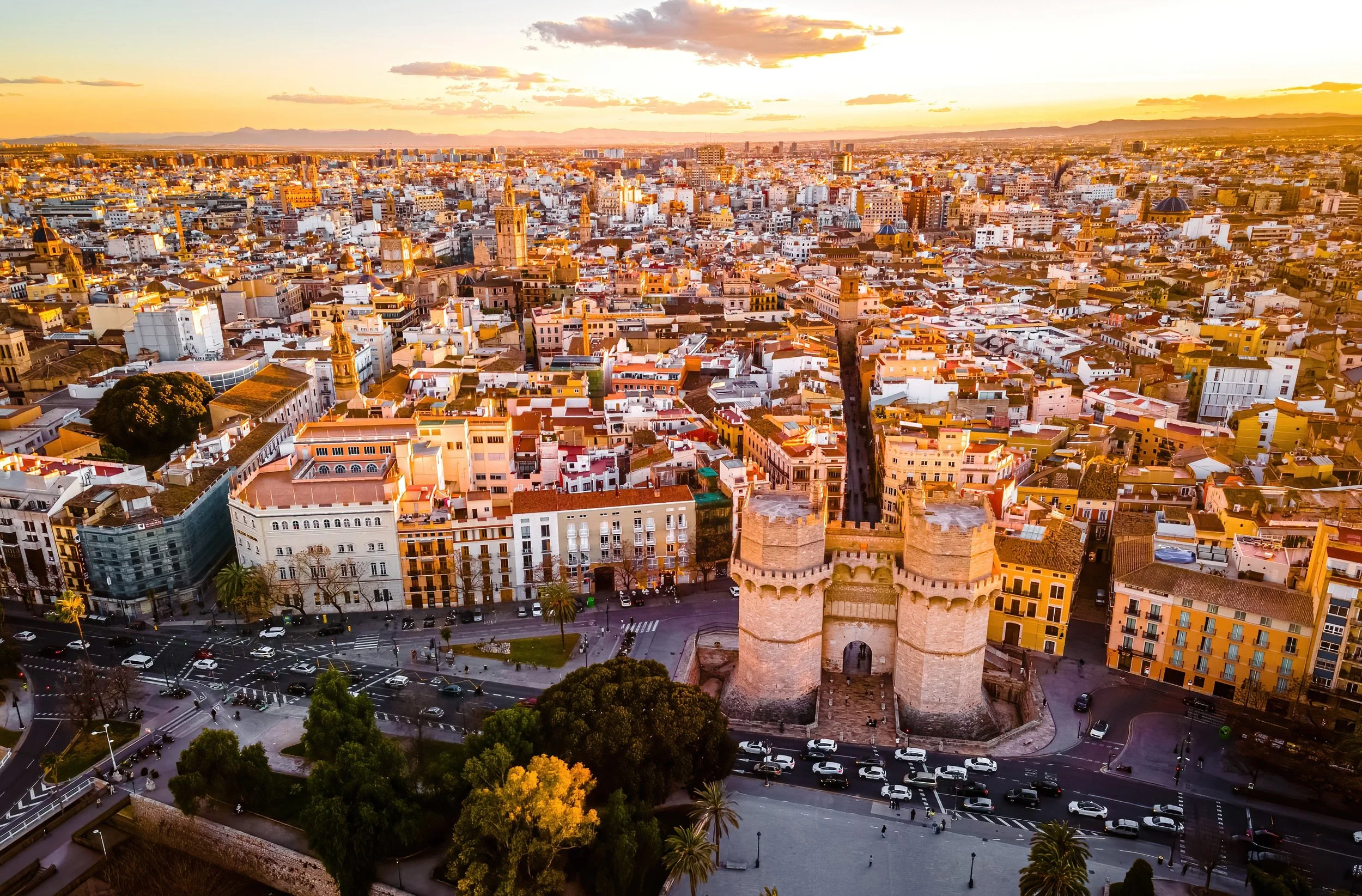 Aerial view of the city of Malaga at sunset, featuring the Alcazaba fortress with its towers in the foreground, surrounded by modern and historic buildings, and a busy street with cars and trees.