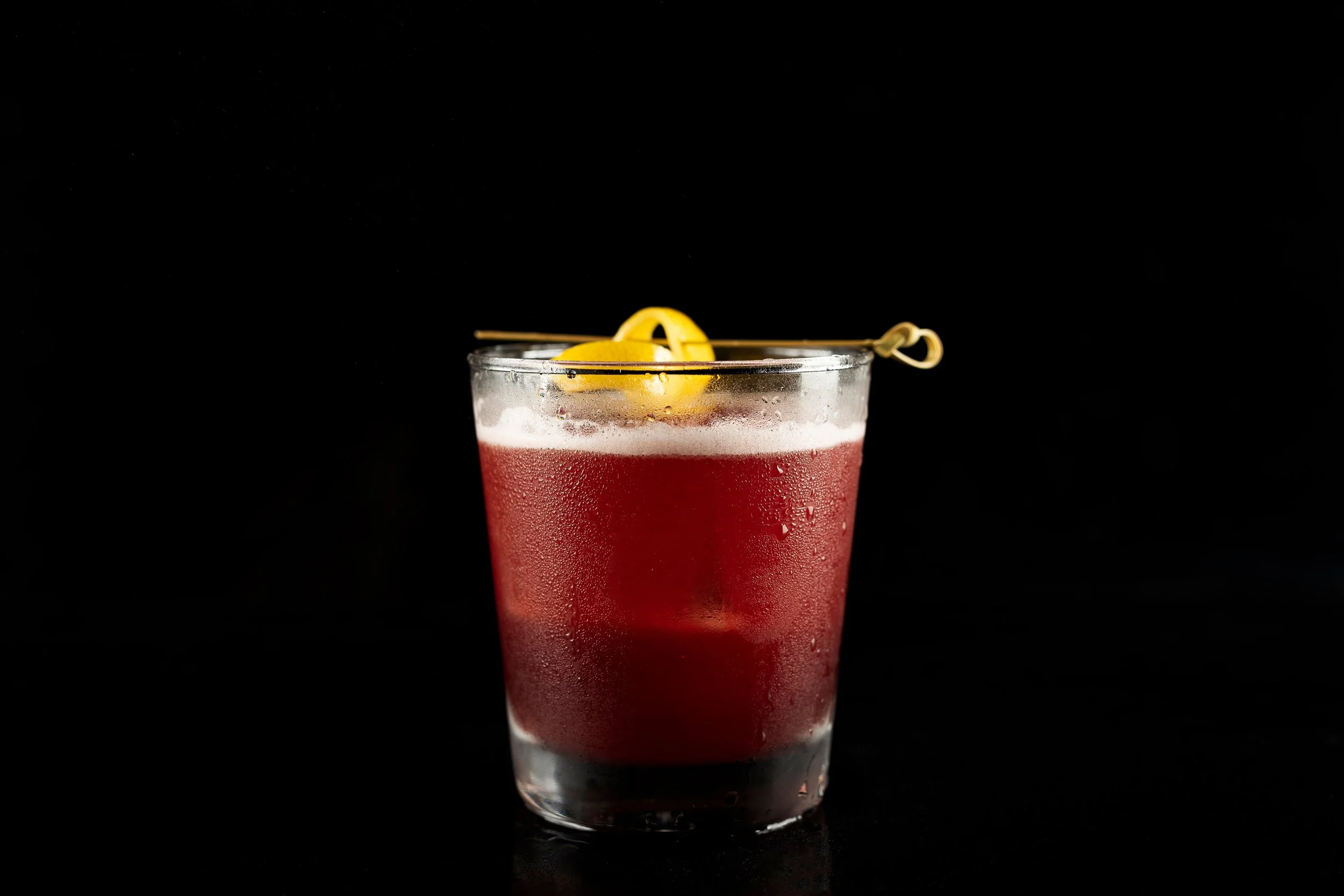 A glass of red cocktail with condensation, garnished with a lemon peel and a decorative stir stick, against a black background.
