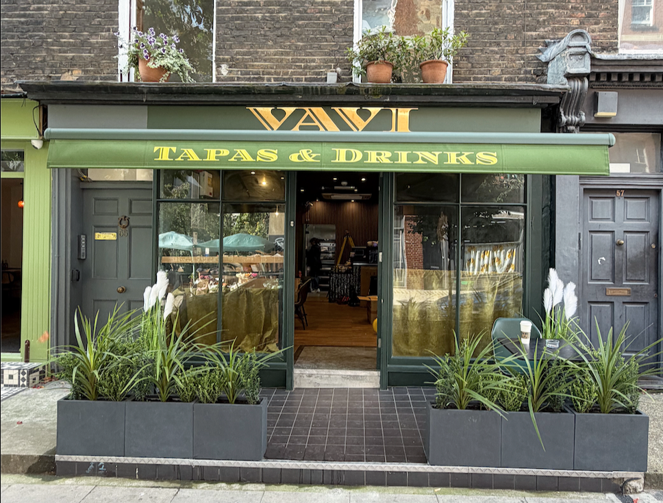 Front view of a tapas and drinks restaurant with a green awning, large glass windows, gray doors, and potted plants outside.