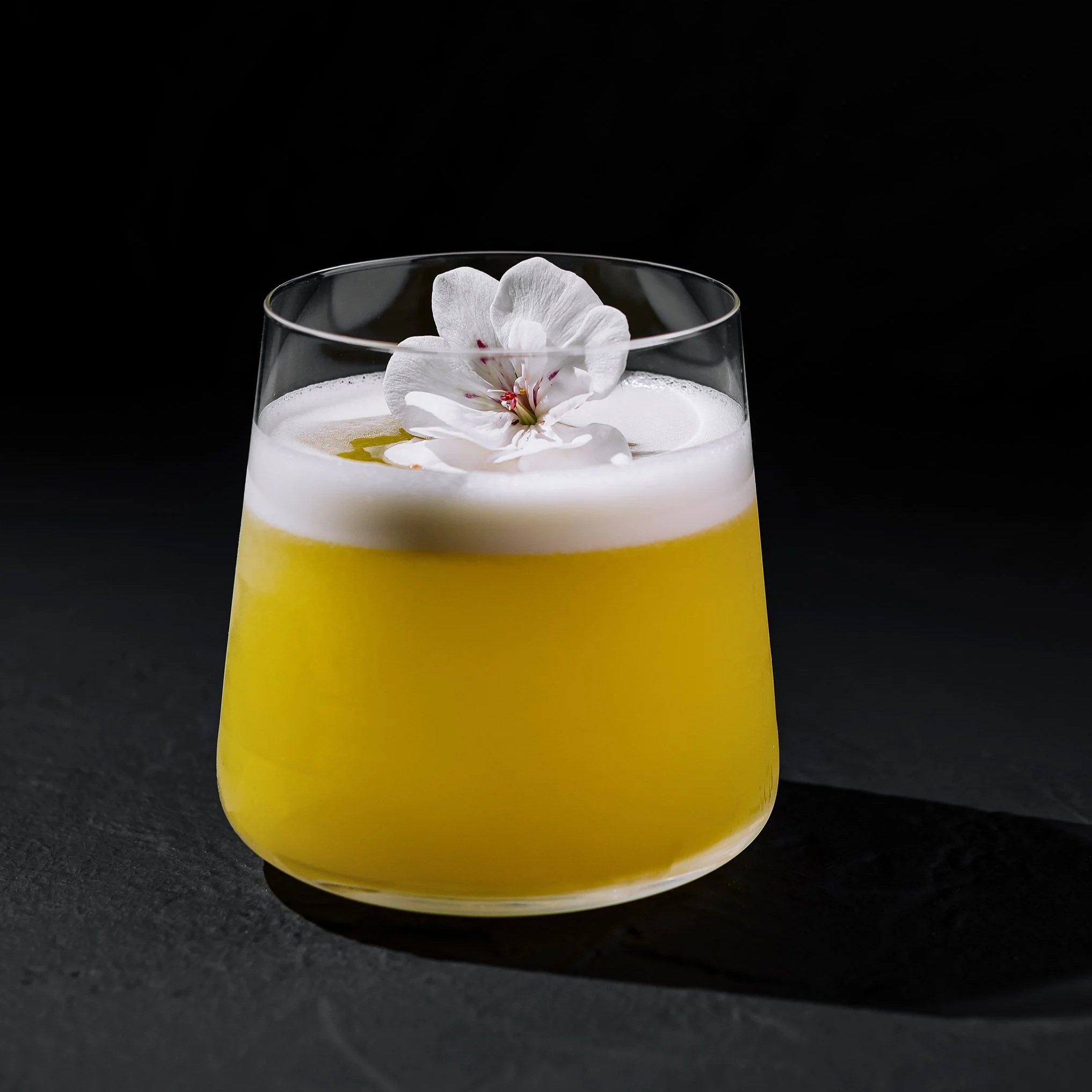 A yellow cocktail in a clear glass with a frothy top, garnished with a white flower on the foam, against a black background.