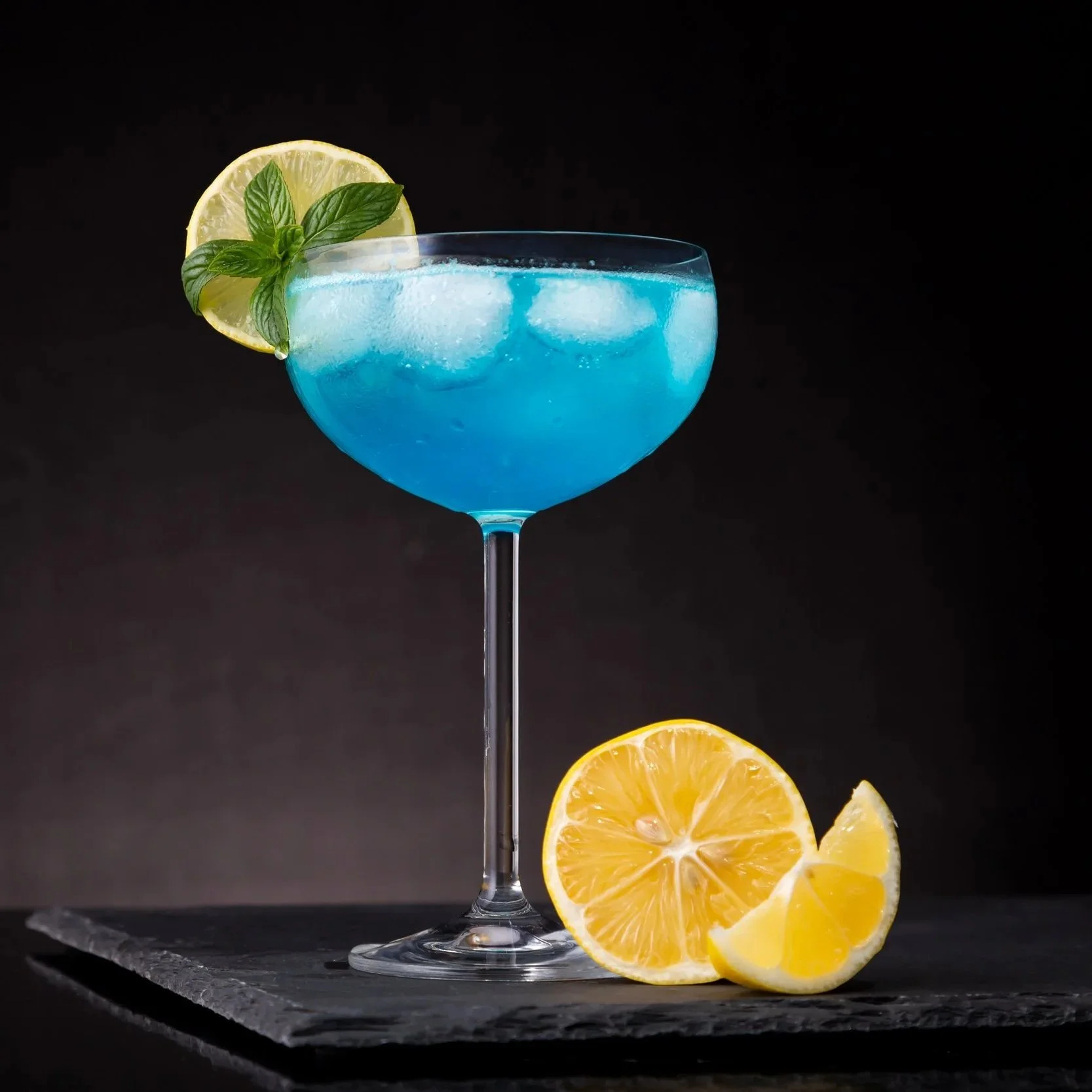Blue cocktail with ice cubes in a coupe glass garnished with a lemon slice and basil leaves, with lemon wedges on a dark slate surface against a black background.
