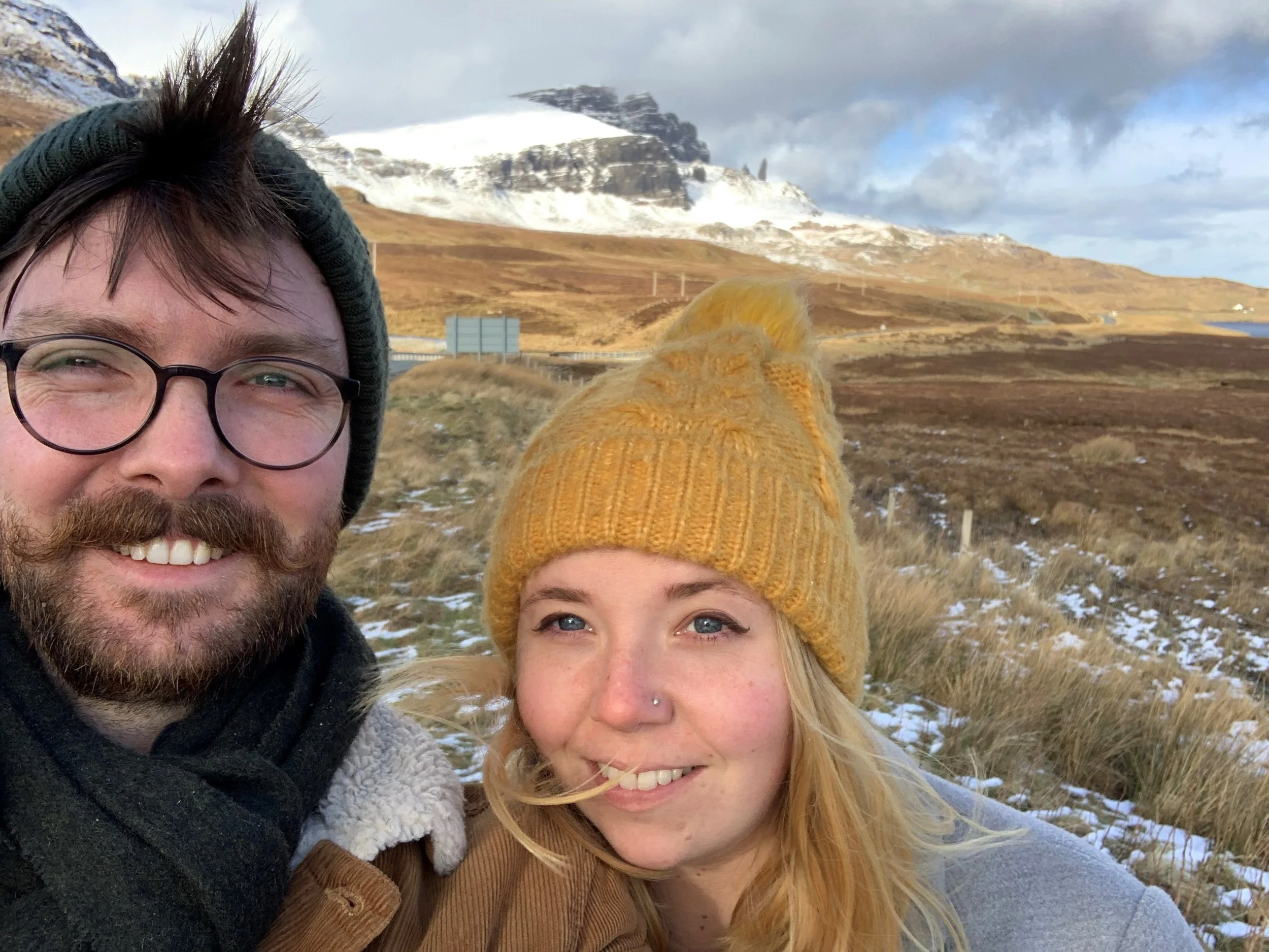 A woman with a yellow knit hat taking a selfie outdoors in a mountainous area with snow patches, hills, and a partly cloudy sky in the background.