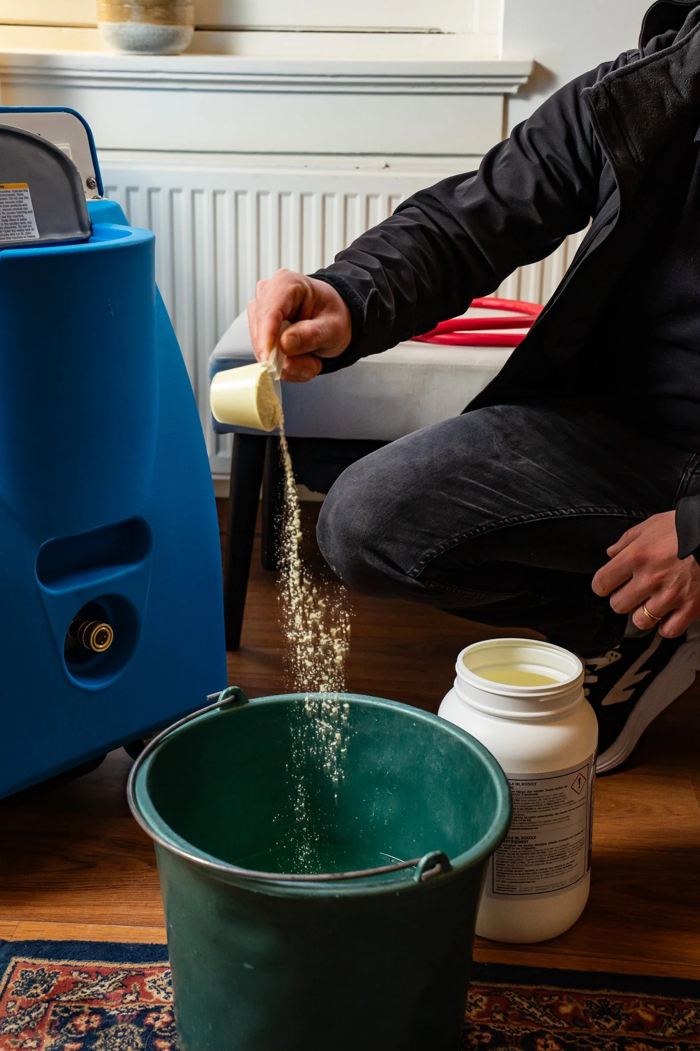 Persoon die poeder uit een siphouer in een groene emmer giet, naast een grote witte container, op een houten vloer met een tapijt. Er staat een blauw apparaat naast, mogelijk een chemisch apparaat of voederautomaat.