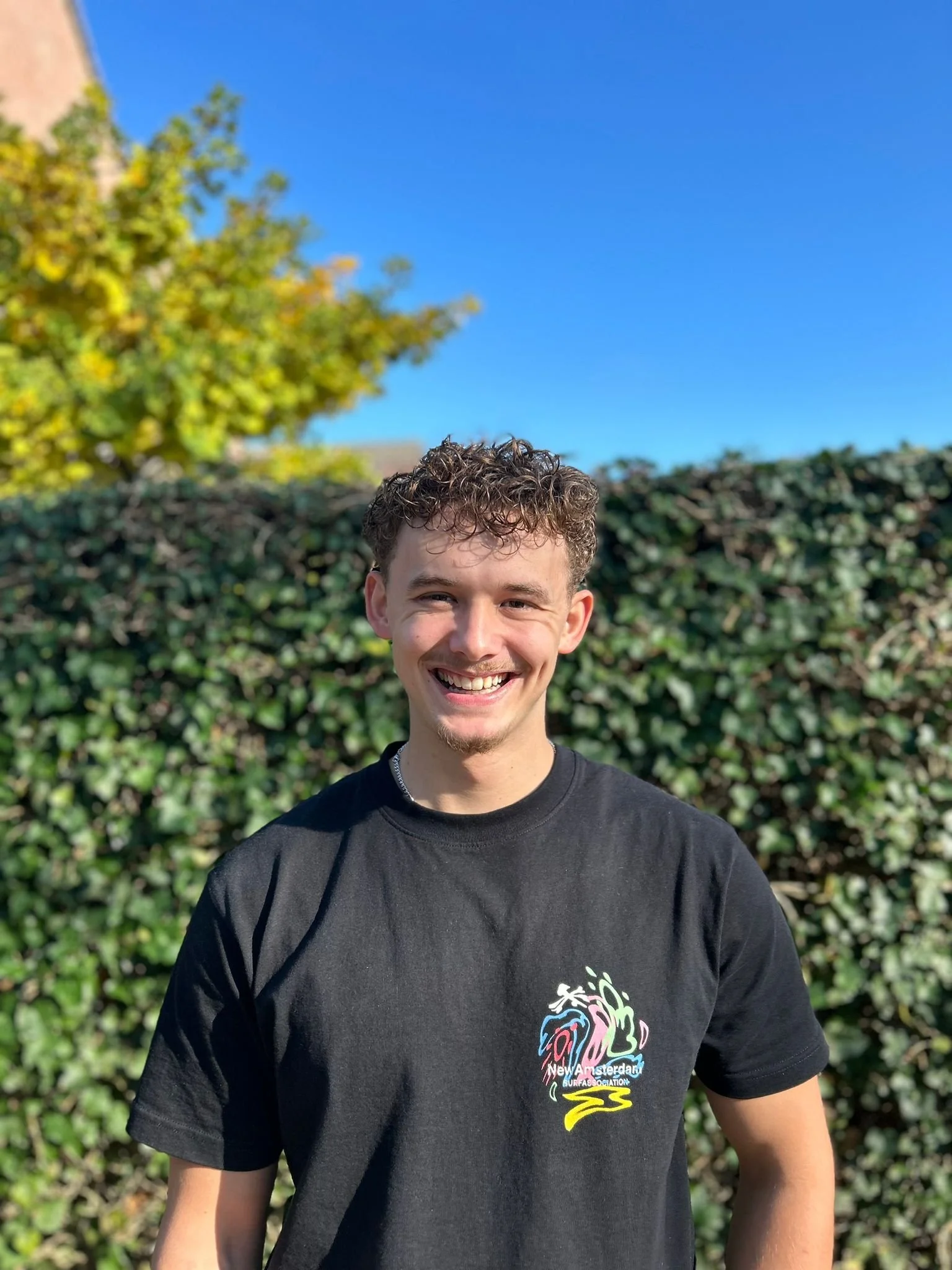 Lachende jonge man met kort, gekruld haar staat voor een heg en een boom, onder een blauwe hemel, draag een zwart T-shirt met kleurrijk logo.