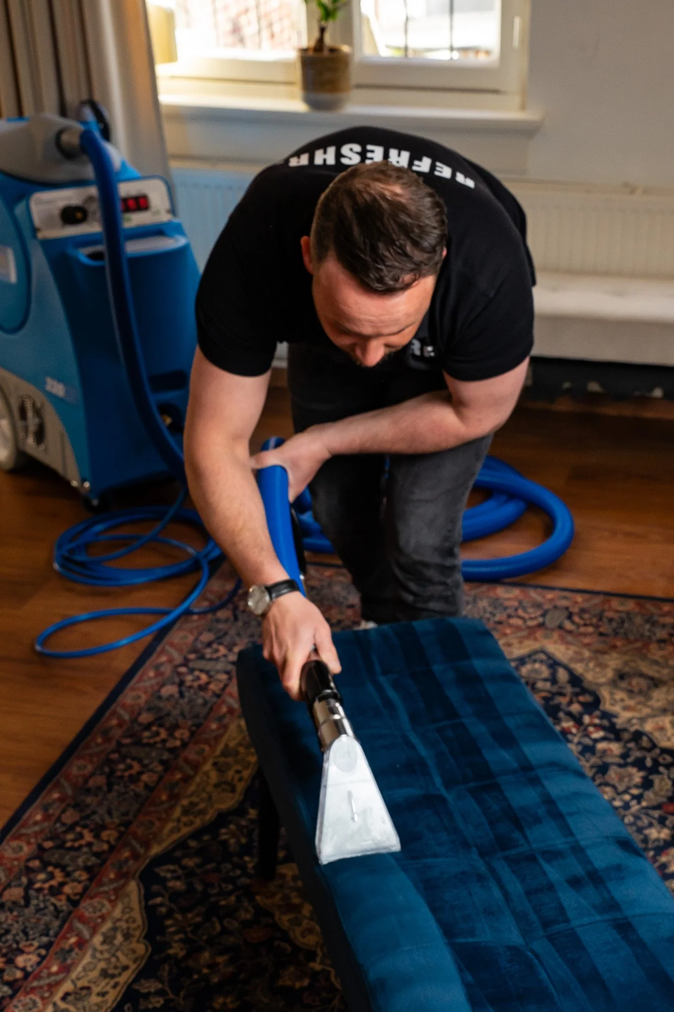 Man ligt op een bank met een stoomzuiger in de hand, reinigt een blauw gestreepte bank. Er staat een blauw machine en slangen op de achtergrond.