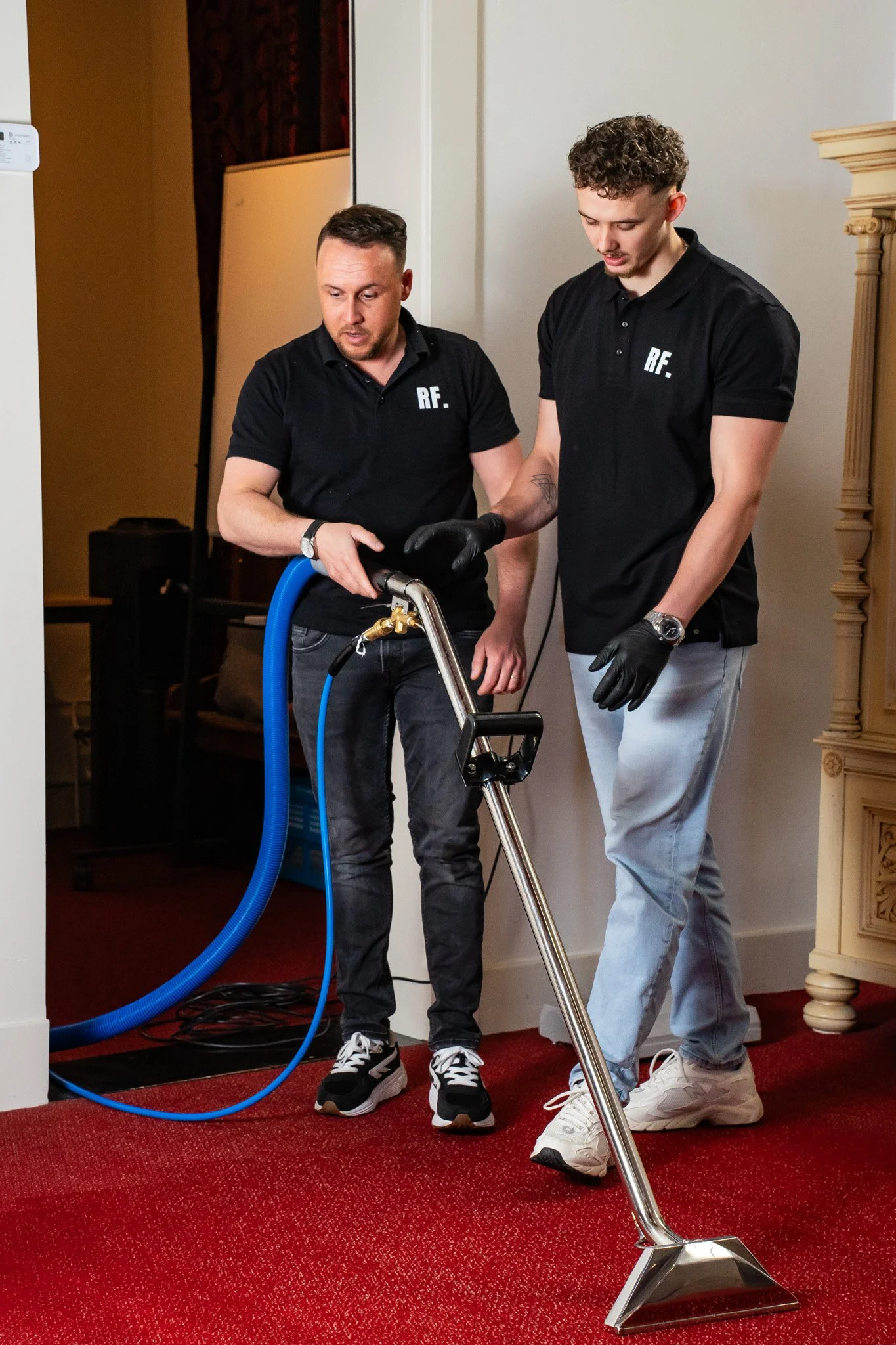 Twee mannen die samen een tapijt reinigen met professionele schoonmaakapparatuur in een interieur met rood tapijt en witte muren.
