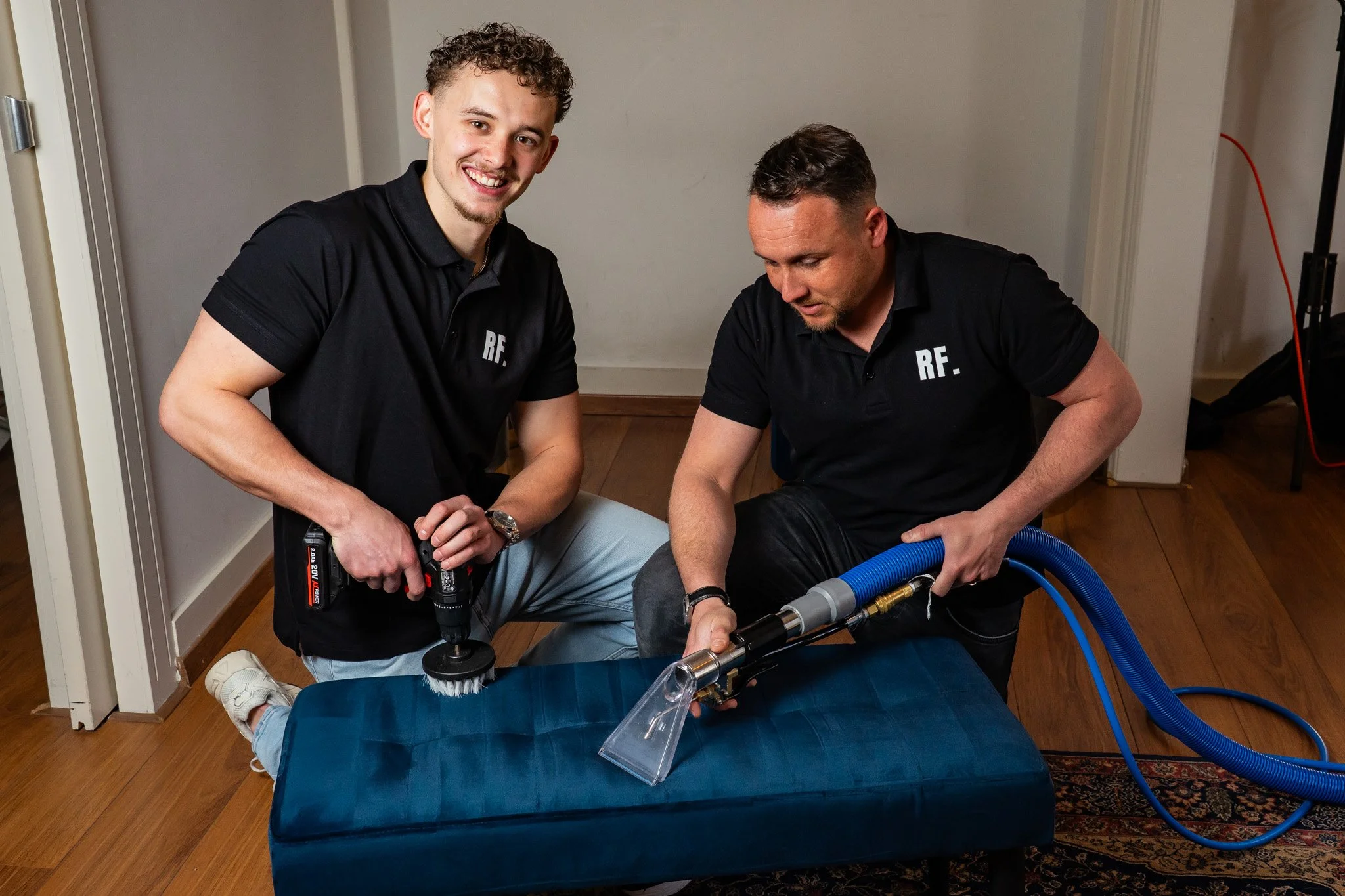 Twee mannen in zwart polo met 'RF' op de borst, gebruikmakend van schoonmaakapparatuur op een blauwe bank. Eén man gebruikt een elektrische polijstmachine, de andere een stofzuiger met een mondstuk, op een houten vloer in een kamer.
