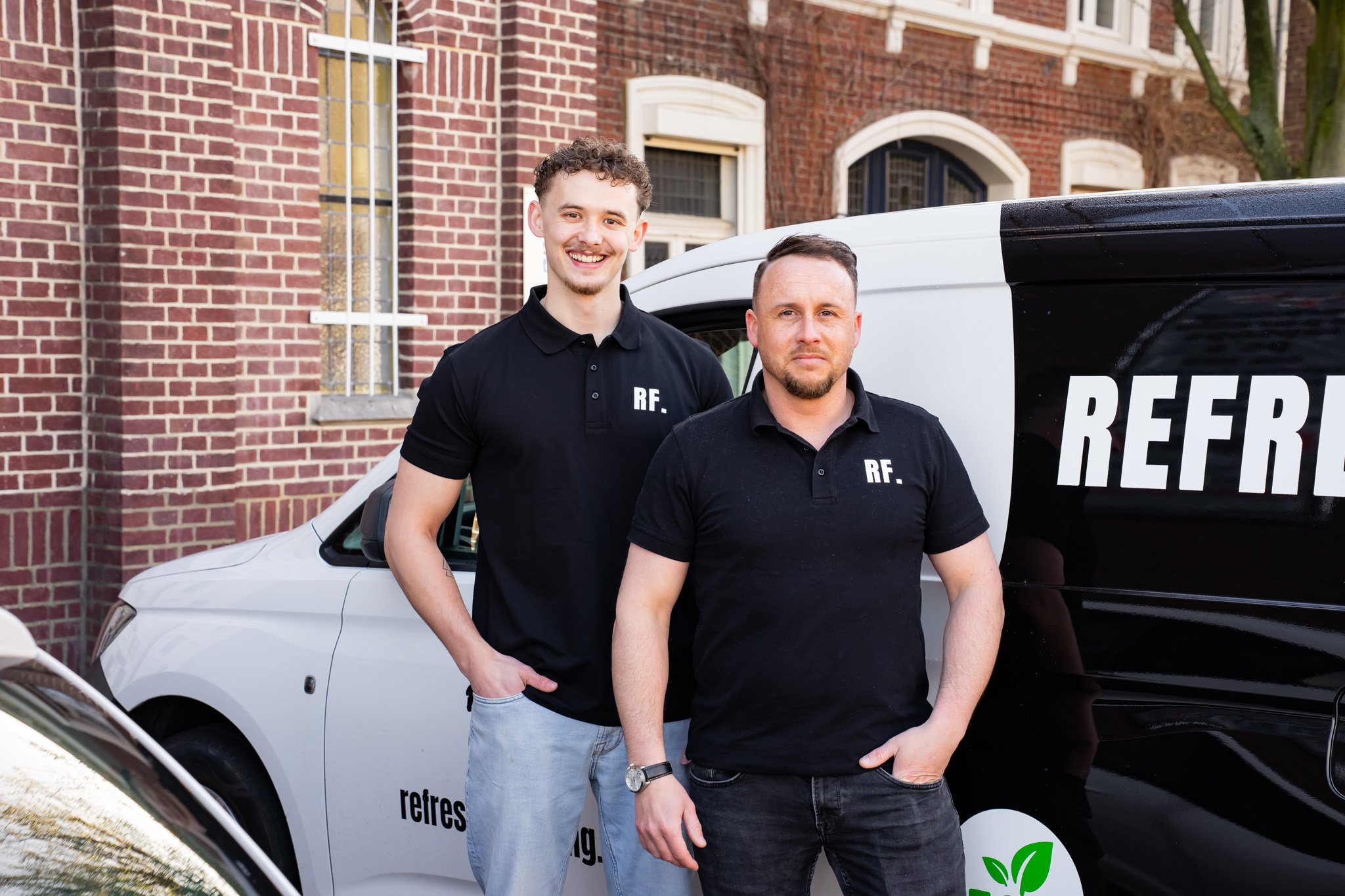Twee mannen in zwart shirt met 'RF.' tekst, staan voor een witte en zwarte bedrijfsbus met 'refres...' tekst, buiten bij een oude bakstenen gebouw.