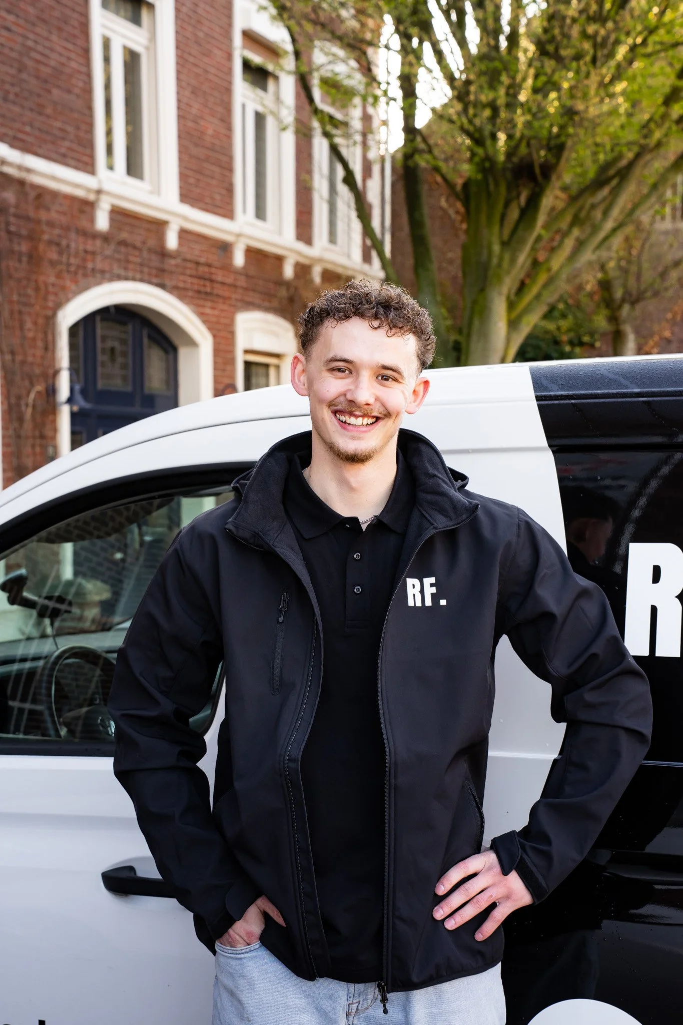 Lachende jonge man met kort, gekruld haar staat voor een heg en een boom, onder een blauwe hemel, draag een zwart T-shirt met kleurrijk logo.