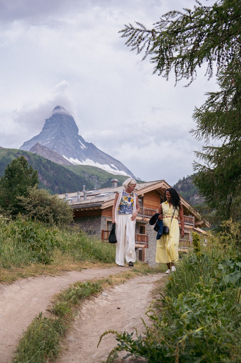 Two women walking on a dirt path in a mountainous area with lush greenery, a large cabin, and a snow-capped mountain in the background.
