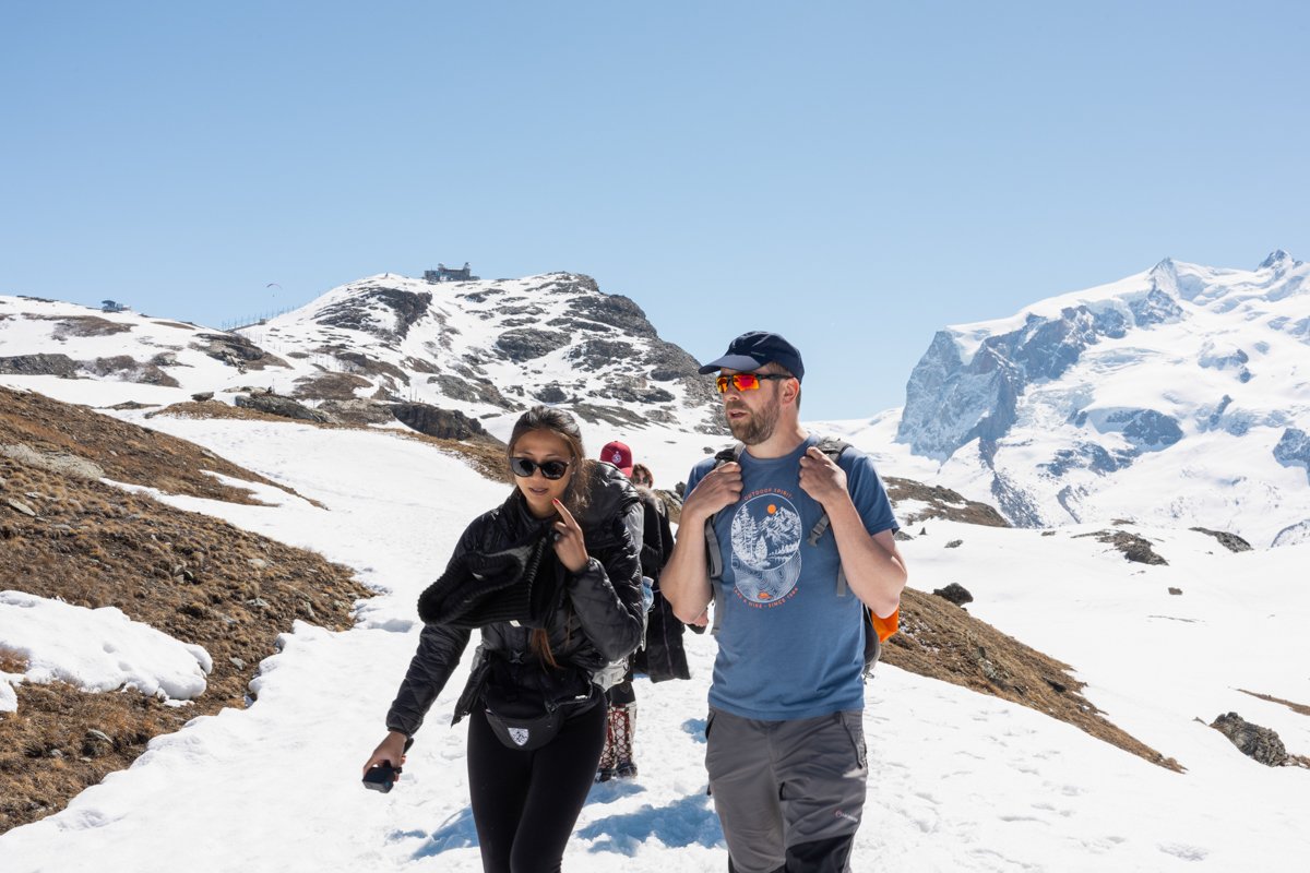 Two hikers walking on a snow-covered mountain trail with snow-capped peaks in the background on a clear, sunny day.