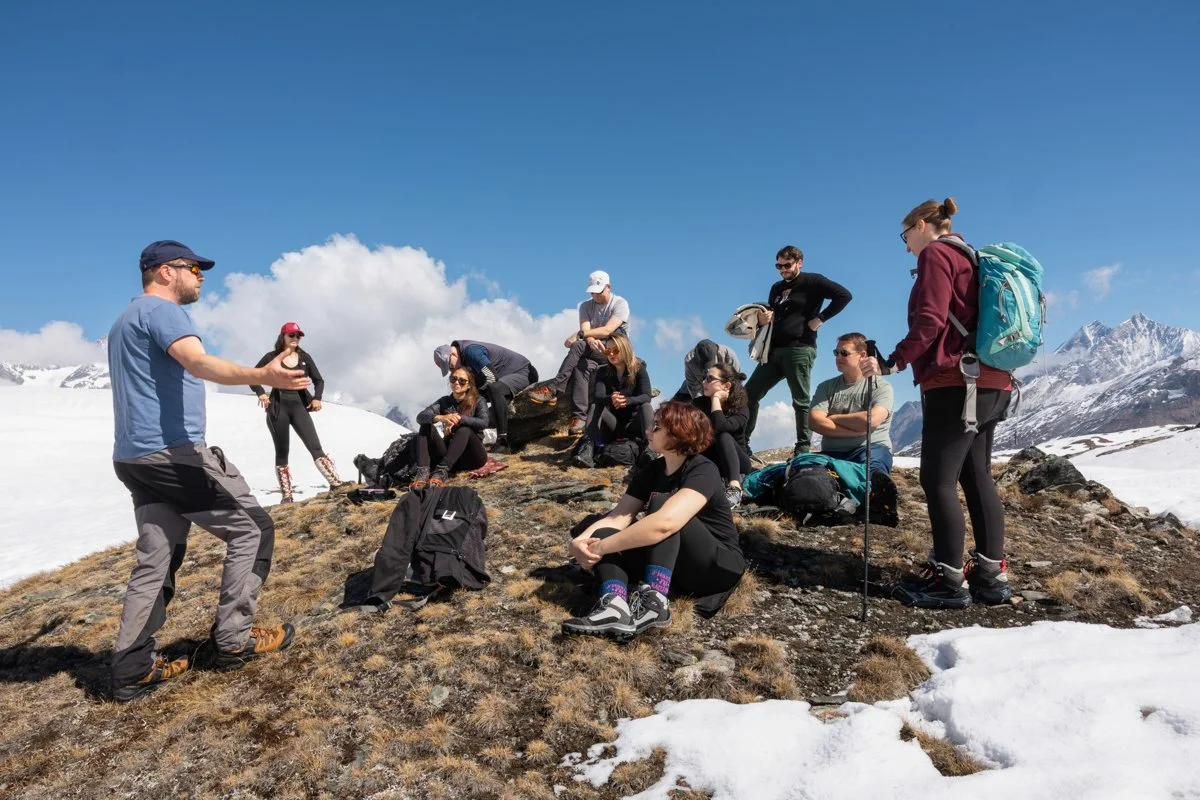 Group of hikers resting on a snowy mountain trail while a guide explains something.