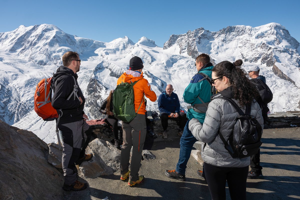 A group of hikers taking a break on a mountain overlook with snow-capped peaks in the background.