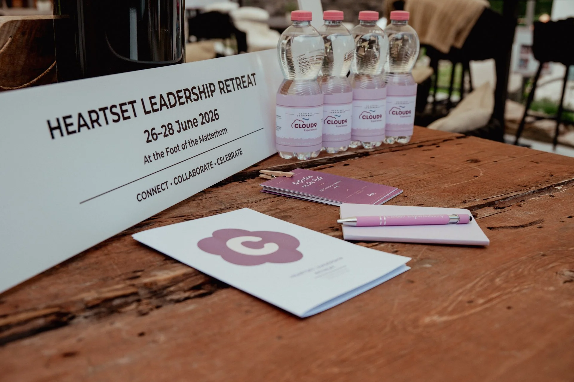 A wooden table with conference materials including a sign for Heartset Leadership Retreat, five water bottles with pink labels and caps, a small purple booklet, a pink pen, and some paper with a purple flower logo.