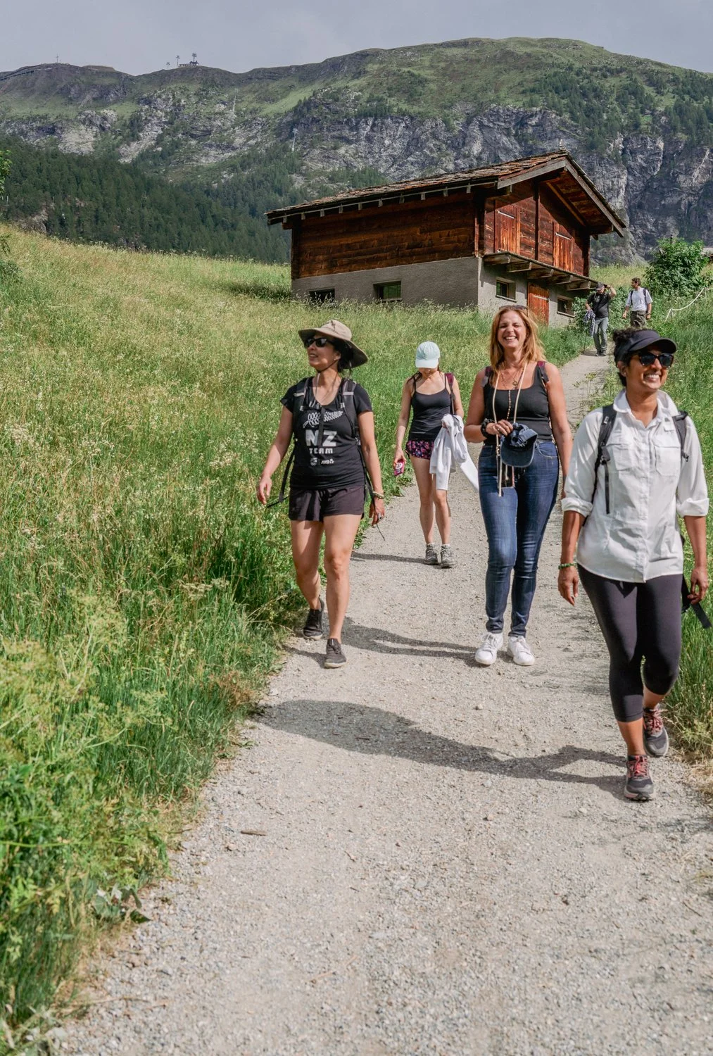 Five women hiking on a dirt trail in a green mountainous landscape with a wooden house in the background.