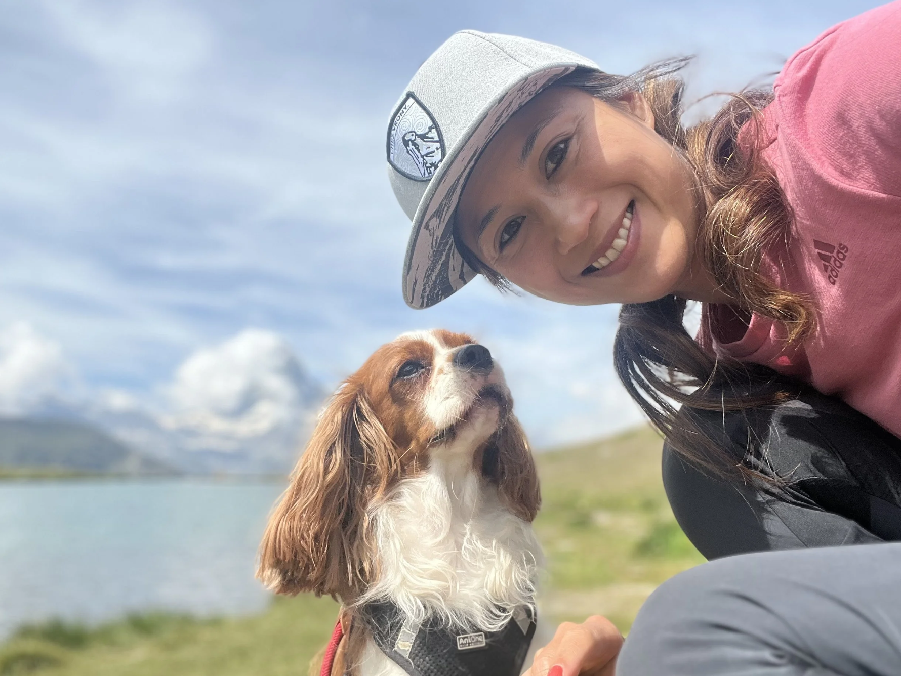 A smiling woman wearing a gray hat and a pink shirt taking a selfie with a brown and white dog outdoors, with a body of water and cloudy sky in the background.