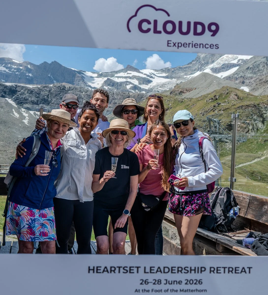 A group of people celebrating at a mountain retreat with snow-capped peaks in the background, holding glasses of champagne, wearing outdoor clothing and hats, at the Heartset Leadership Retreat from June 26-28, 2026, at the foot of the Matterhorn.