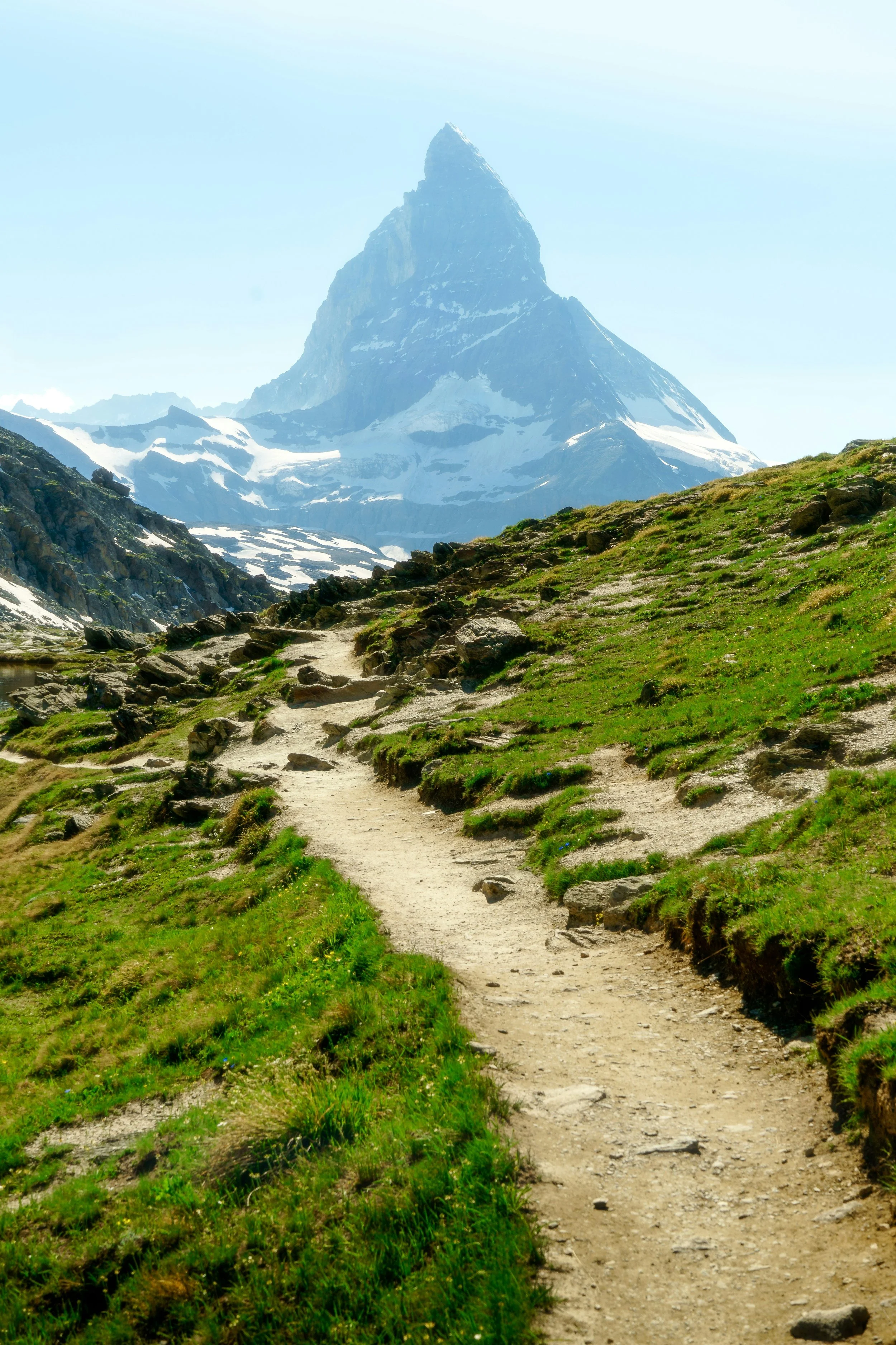 A dirt trail winding through green grass and rocks, leading towards a snow-capped mountain with a pointed peak in the background.