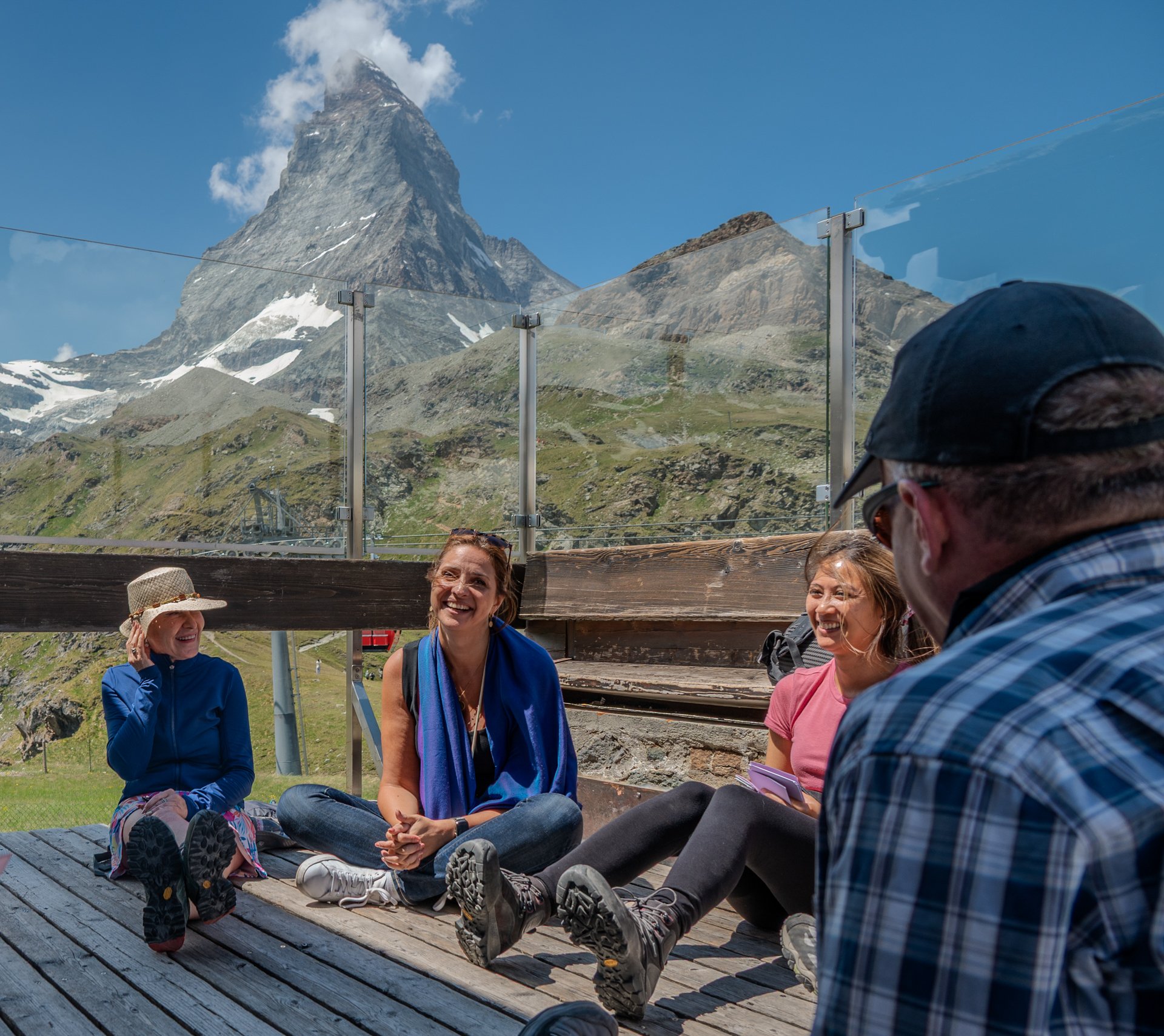 Group of people sitting on a wooden deck outdoors with mountains and blue sky in the background, enjoying a sunny day.
