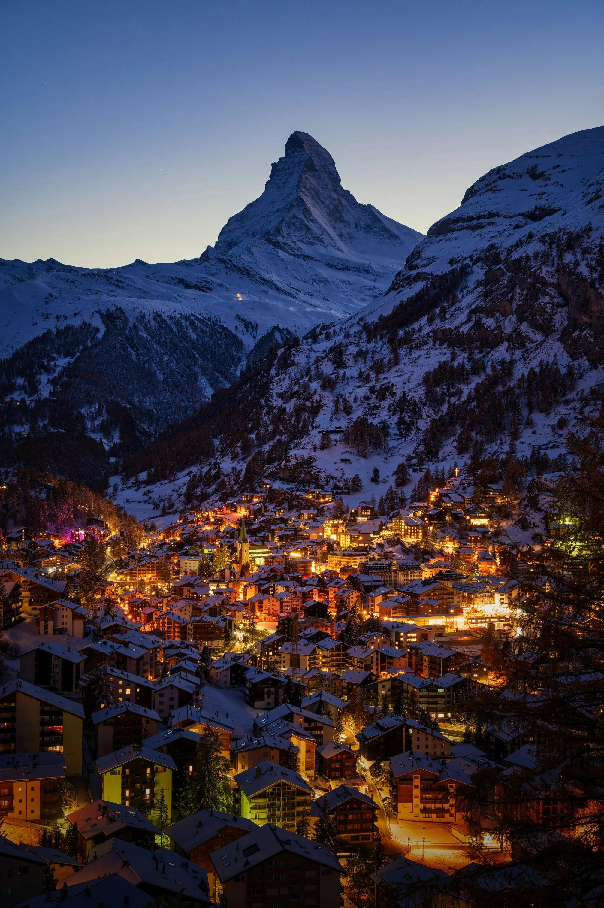 Night view of a snow-covered mountain with a small town at the base illuminated by streetlights and building lights.