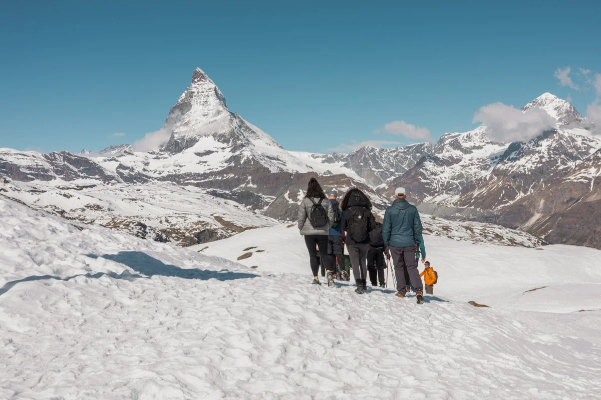 A group of hikers walking on snow-covered terrain in the mountains with snow-capped peaks and clear blue sky in the background.
