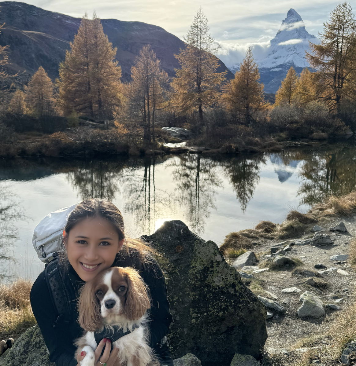 A smiling woman with a dog near a lake in a mountainous landscape with autumn trees and a snow-capped mountain in the background.