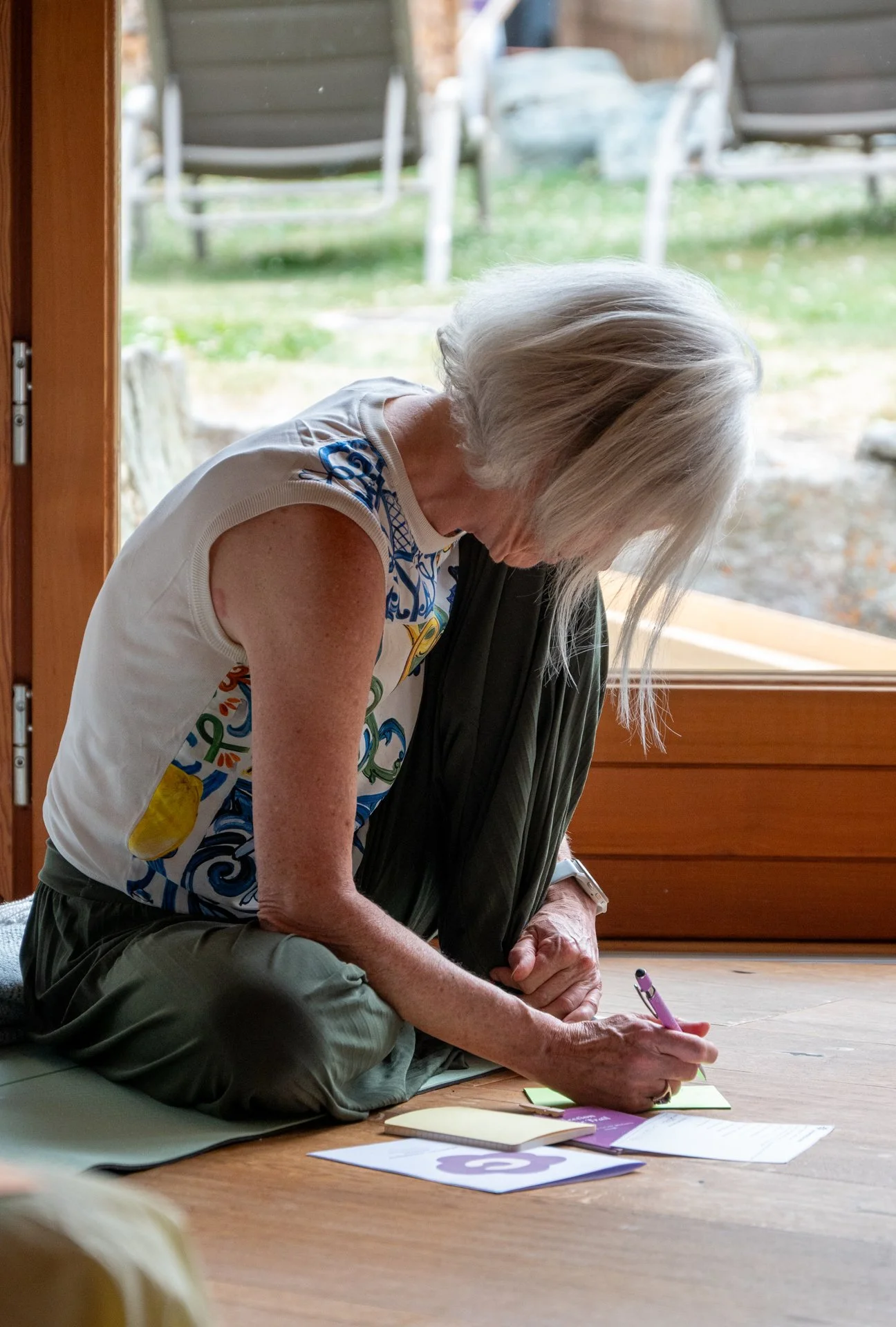 An elderly woman with white hair sitting on a mat on the floor, writing on a notepad with a purple pen, surrounded by papers and notebooks, near a large window with a view of an outdoor yard with benches and rocks.