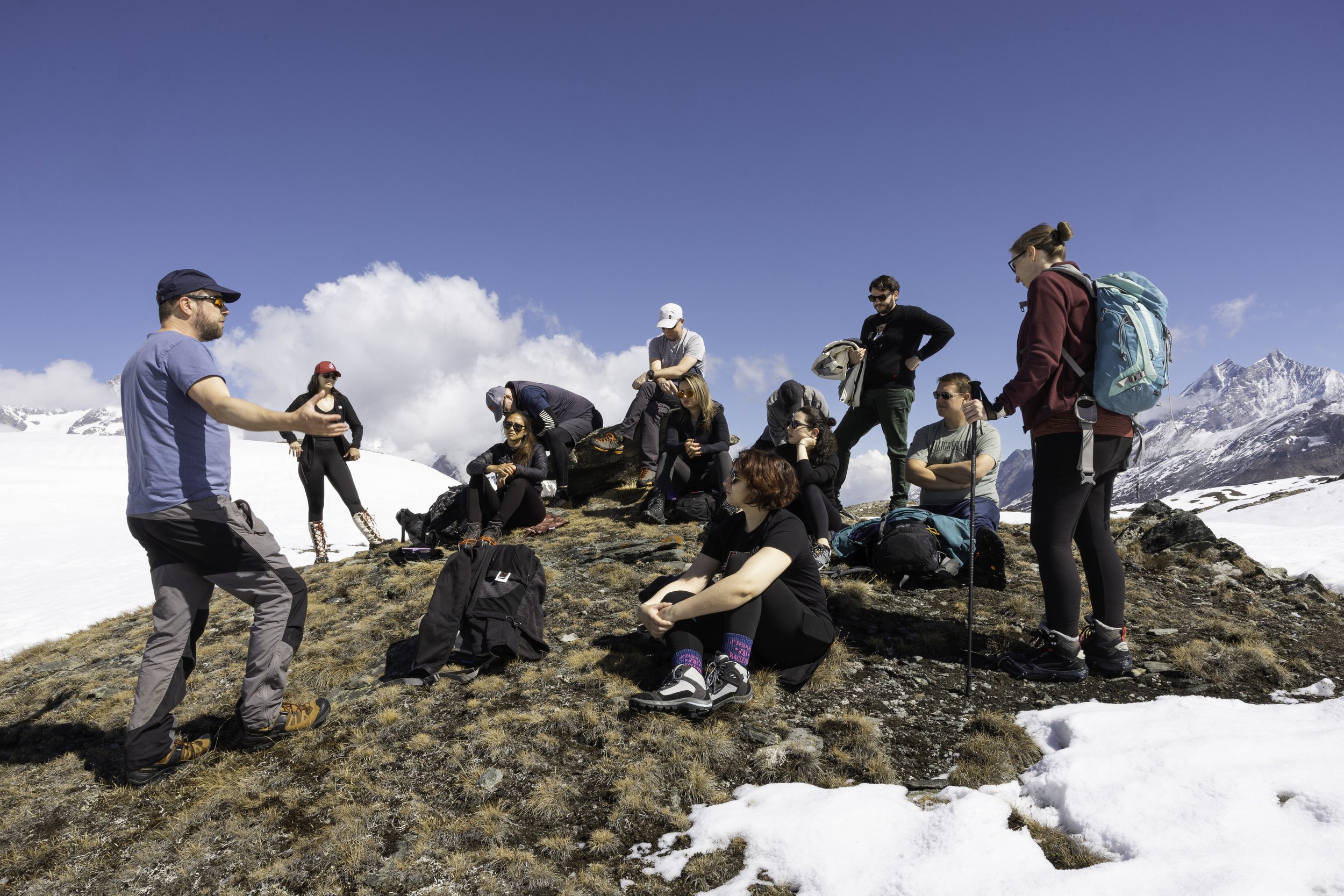 Group of hikers resting on a mountain trail with snow patches, rugged mountains, and a bright blue sky in the background.