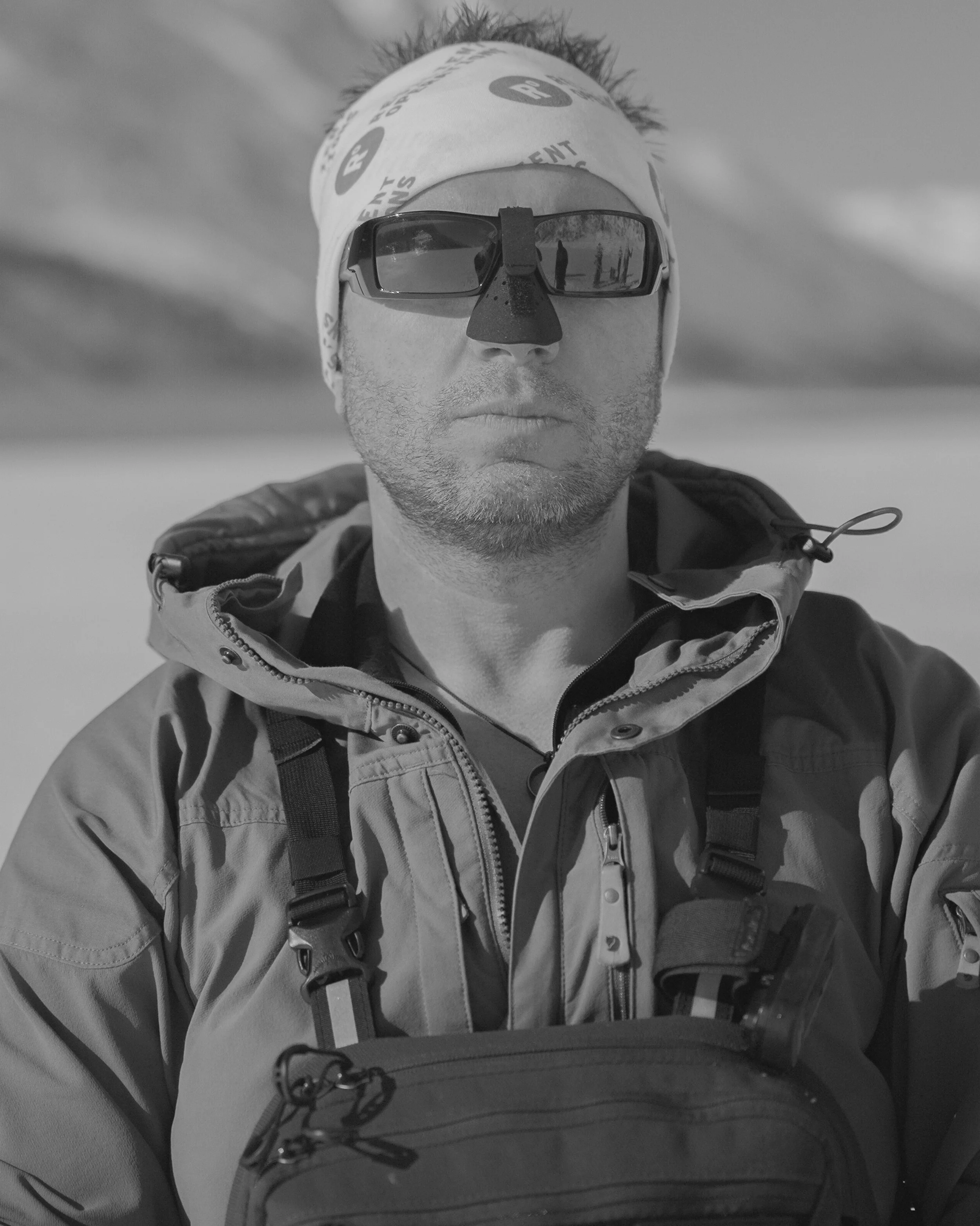 A man wearing sunglasses, a bandana, and a jacket outdoors, with a landscape and cloudy sky in the background.